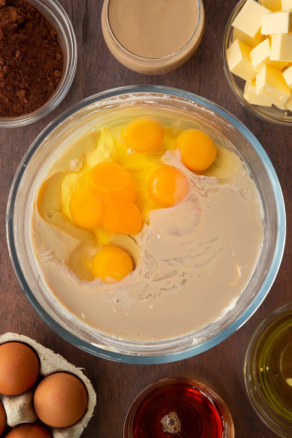Butter, vegetable oil, caster sugar and golden syrup, whisked together in a glass mixing bowl with egg and Baileys on top. Ingredients to make a Baileys cake surround the bowl.