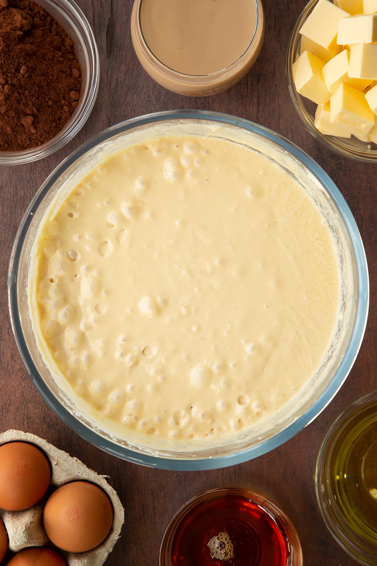 Butter, vegetable oil, caster sugar, golden syrup, egg and Baileys, whisked together in a glass mixing bowl. Ingredients to make a Baileys cake surround the bowl.