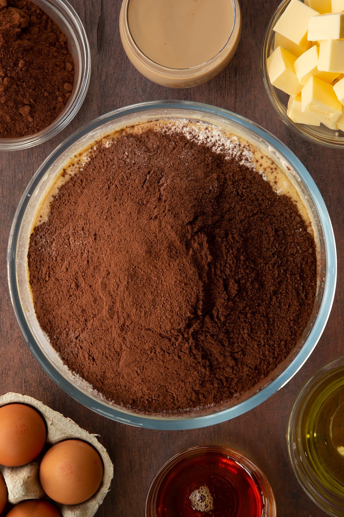 Butter, vegetable oil, caster sugar, golden syrup, egg and Baileys, whisked together in a glass mixing bowl with flour and cocoa on top. Ingredients to make a Baileys cake surround the bowl.