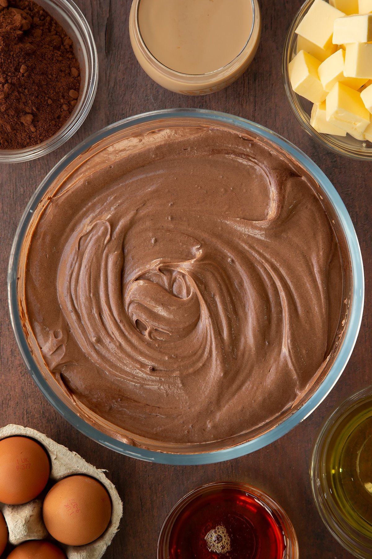 Chocolate Baileys cake batter in a glass mixing bowl. Ingredients to make a Baileys cake surround the bowl.