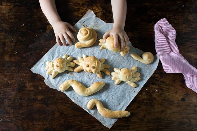 Bread in the shape of spiders and snakes on baking paper. A child's hands reach for the bread.