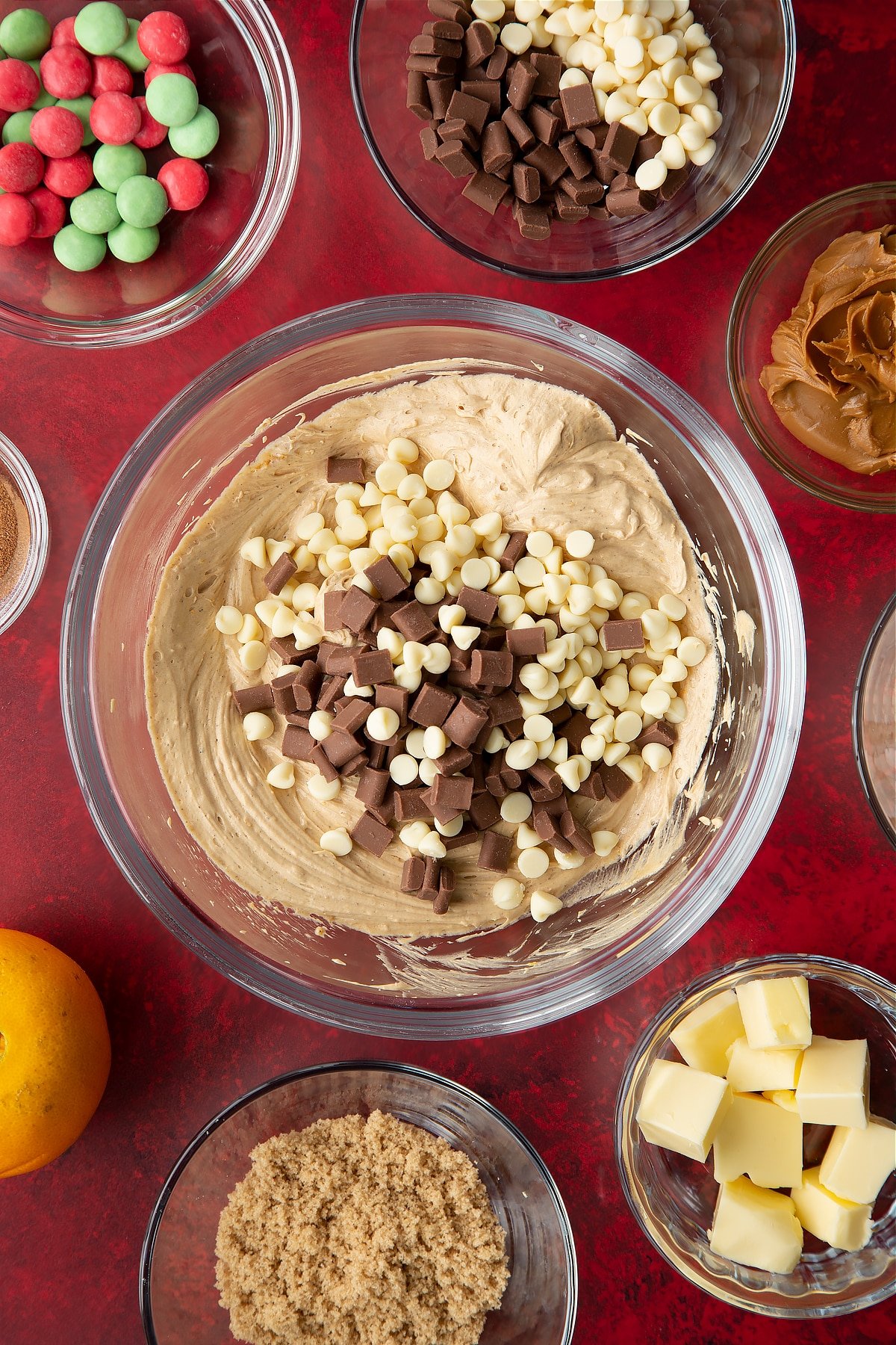 Butter, cream cheese, Biscoff spread, sugar, orange zest and cinnamon, whisked together in a mixing bowl with white and milk chocolate chips on top. Ingredients to make Christmas cookie dip surround the bowl.