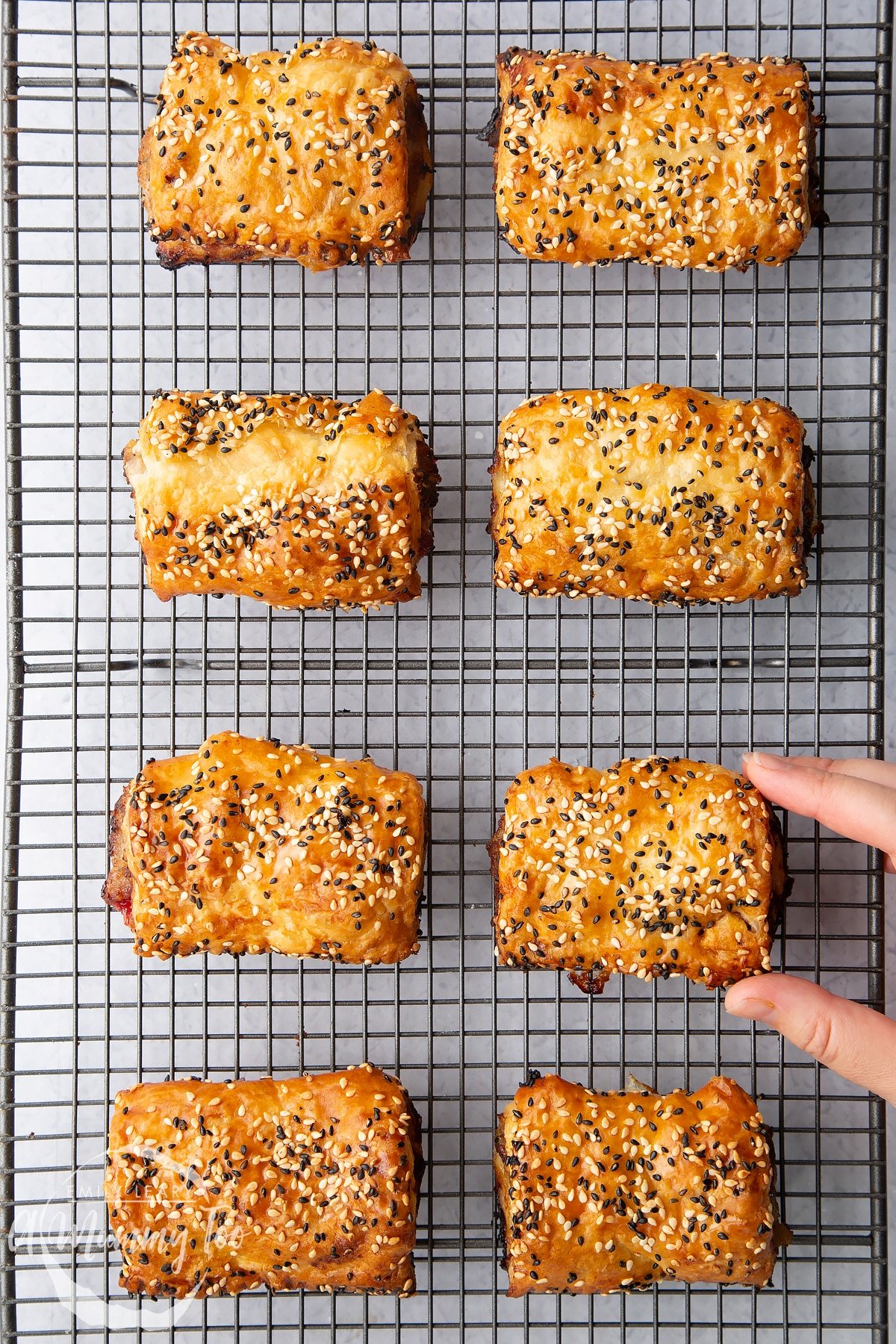 Festive sausage rolls on a wire cooling rack. A hand reaches for one.