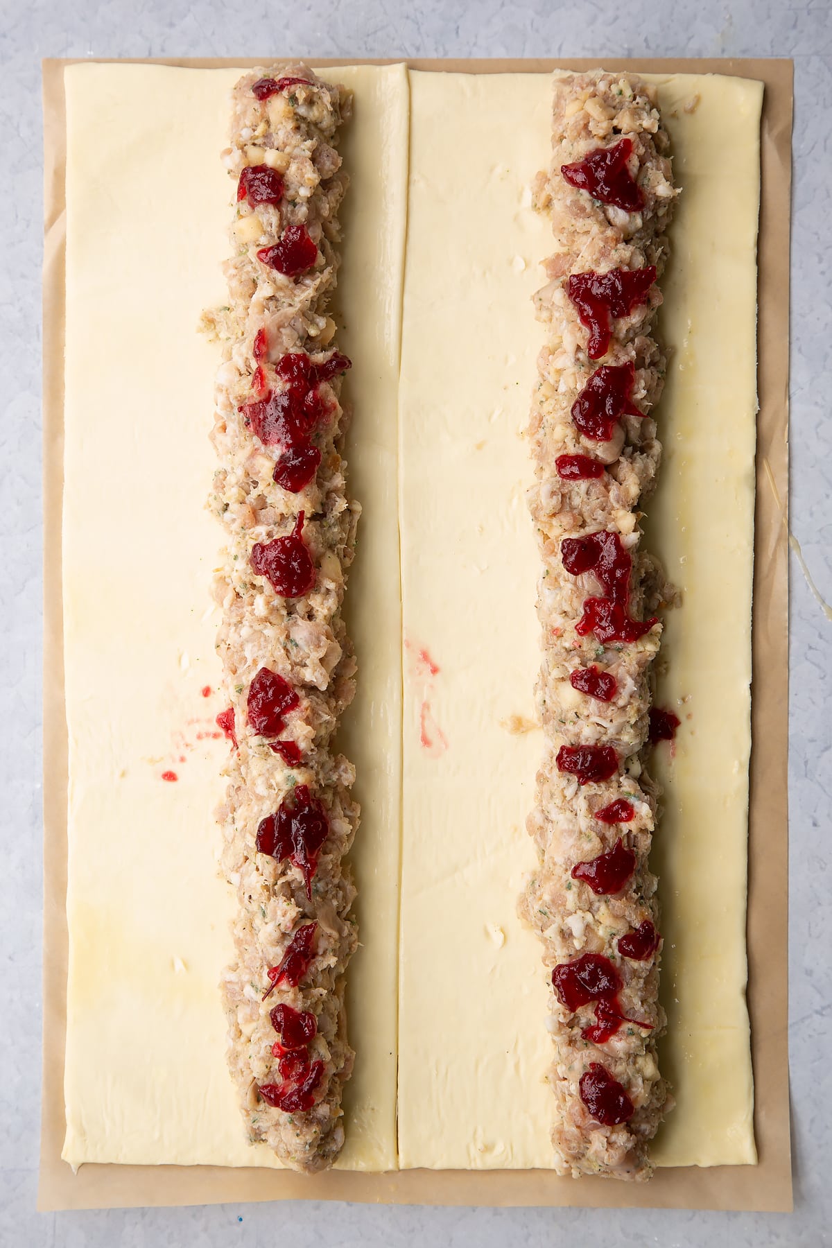 A sheet of puff pastry cut in half lengthways. Each half is egg washed and has a length of festive sausage meat down the middle, dotted with cranberry sauce.