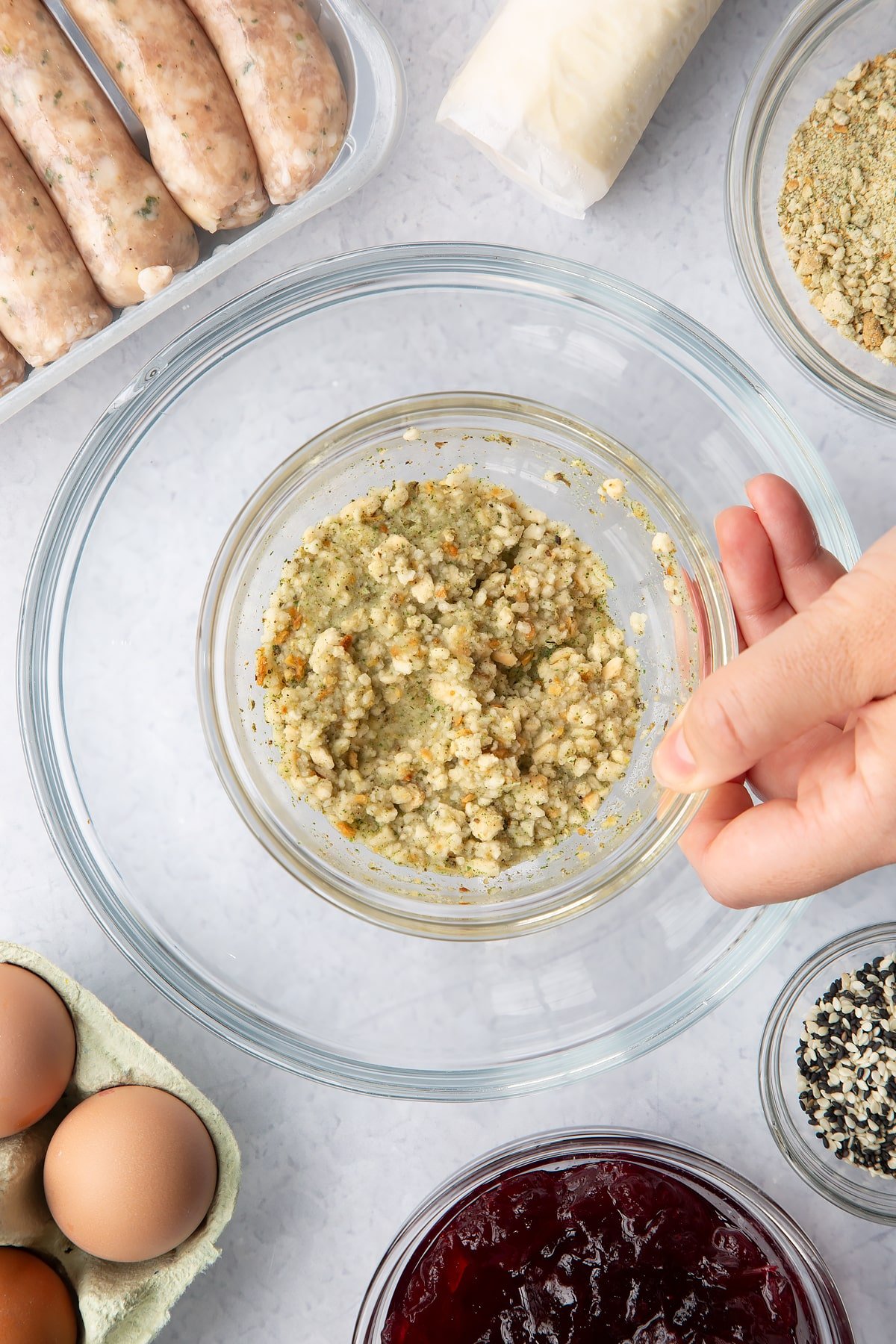 A hand holding wet stuffing in a small bowl above ingredients to make a festive sausage roll recipe