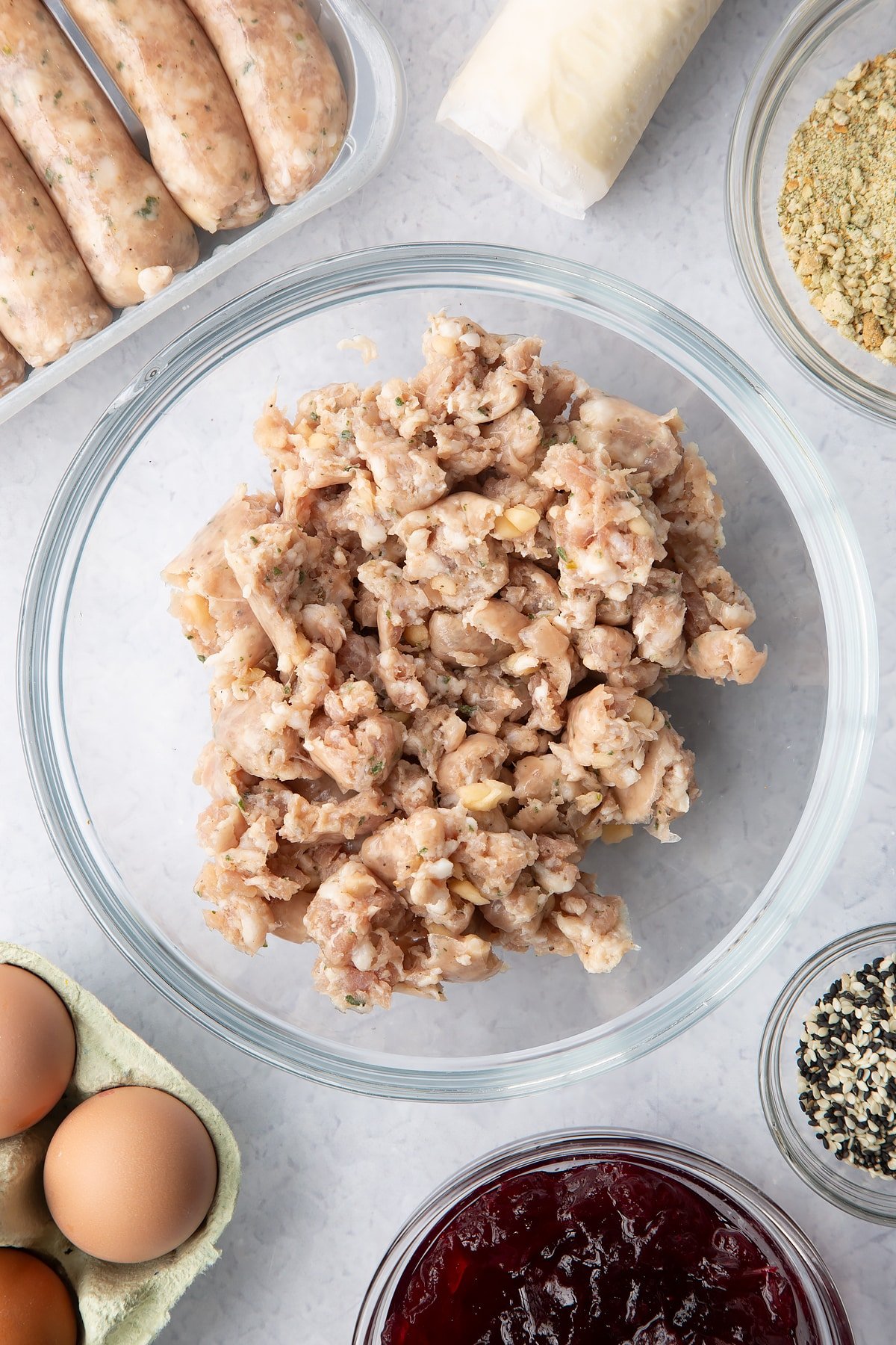 Sausage meat in a glass mixing bowl. Ingredients to make a festive sausage roll recipe surround the bow.