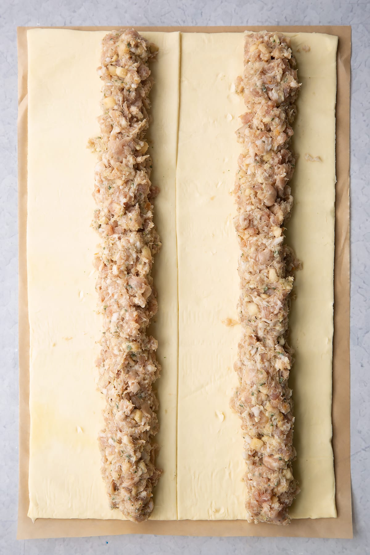 A sheet of puff pastry cut in half lengthways. Each half has a length of festive sausage meat down the middle.