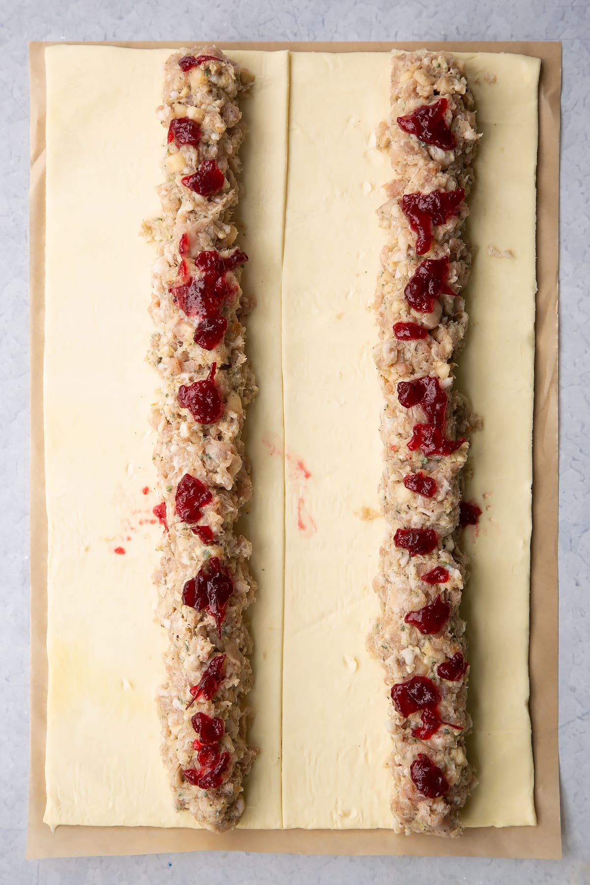 A sheet of puff pastry cut in half lengthways. Each half has a length of festive sausage meat down the middle, dotted with cranberry sauce.