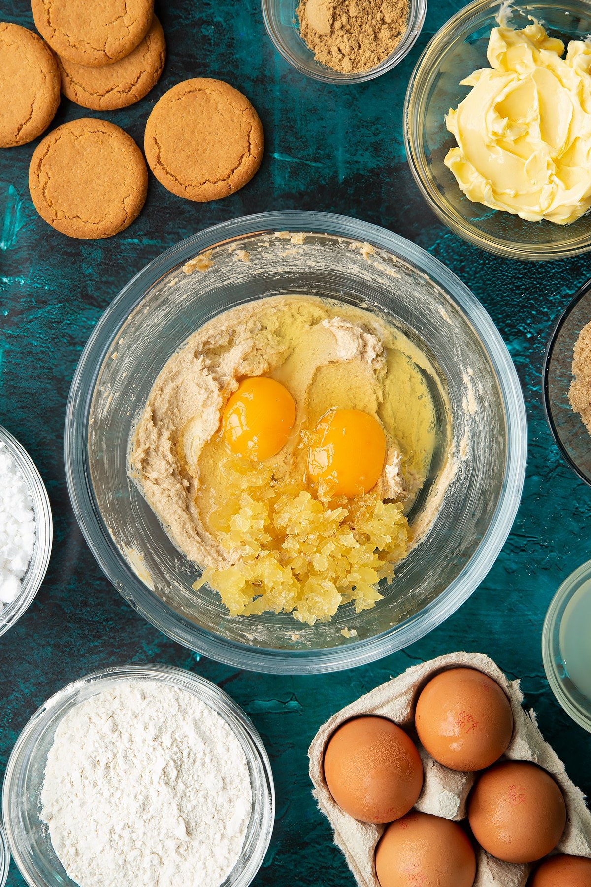 Butter and brown sugar beaten together in a glass mixing bowl with eggs, vanilla and chopped stem ginger on top. Ingredients to make gingerbread donuts surround the bowl.