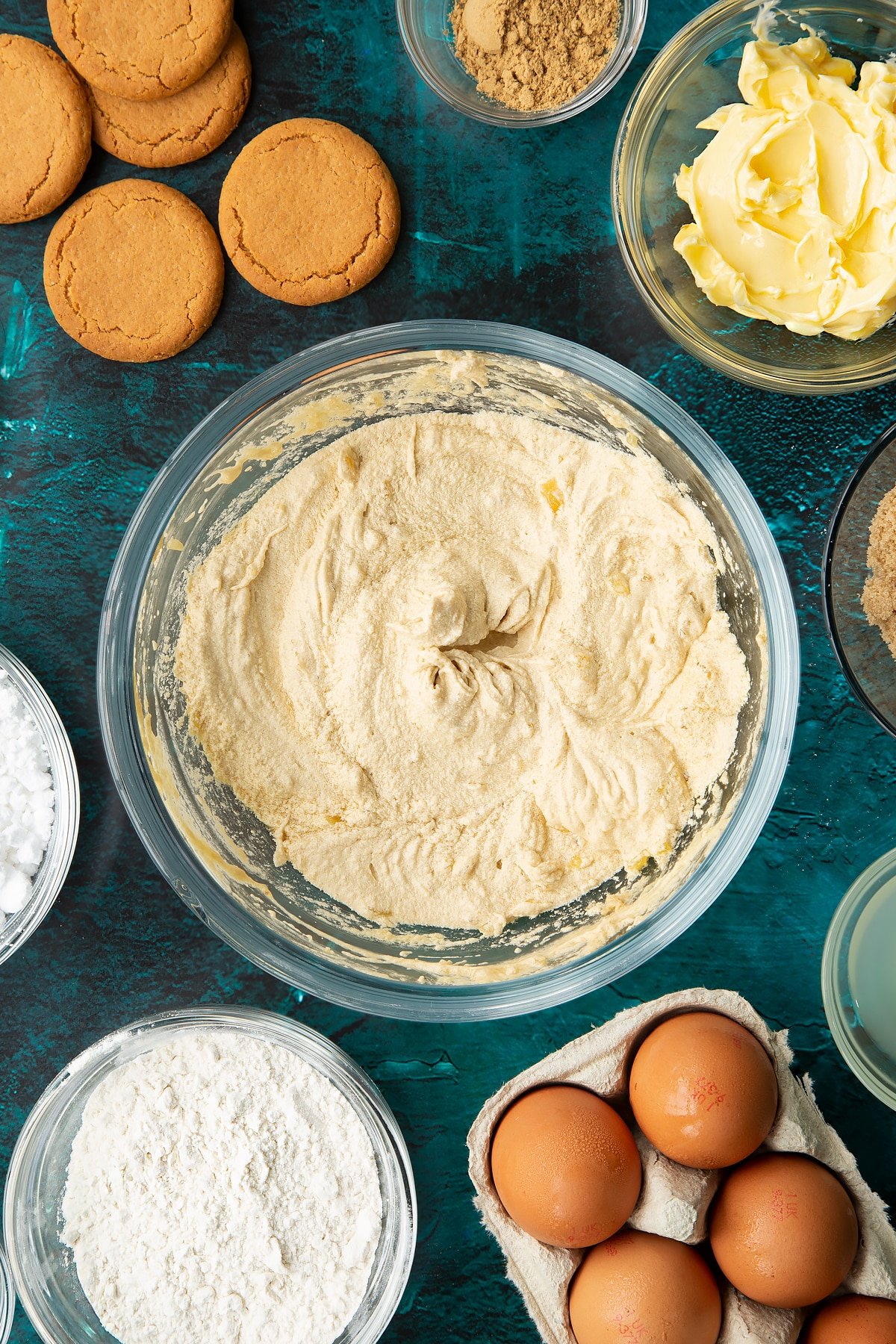 Butter, brown sugar, eggs, vanilla and chopped stem ginger beaten together in a glass mixing bowl. Ingredients to make gingerbread donuts surround the bowl.