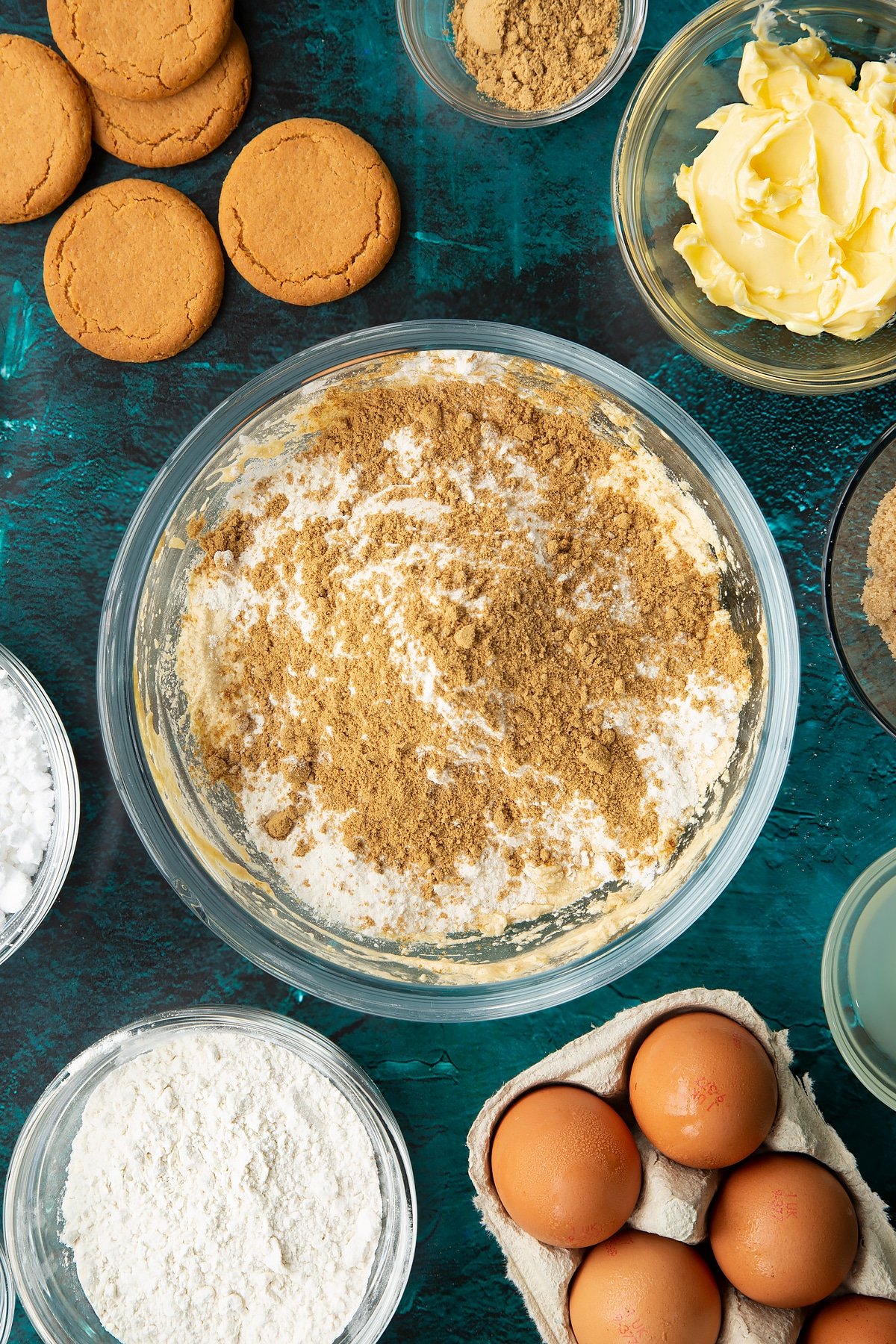 Butter, brown sugar, eggs, vanilla and chopped stem ginger beaten together in a glass mixing bowl with flour, baking powder and ginger on top. Ingredients to make gingerbread donuts surround the bowl.