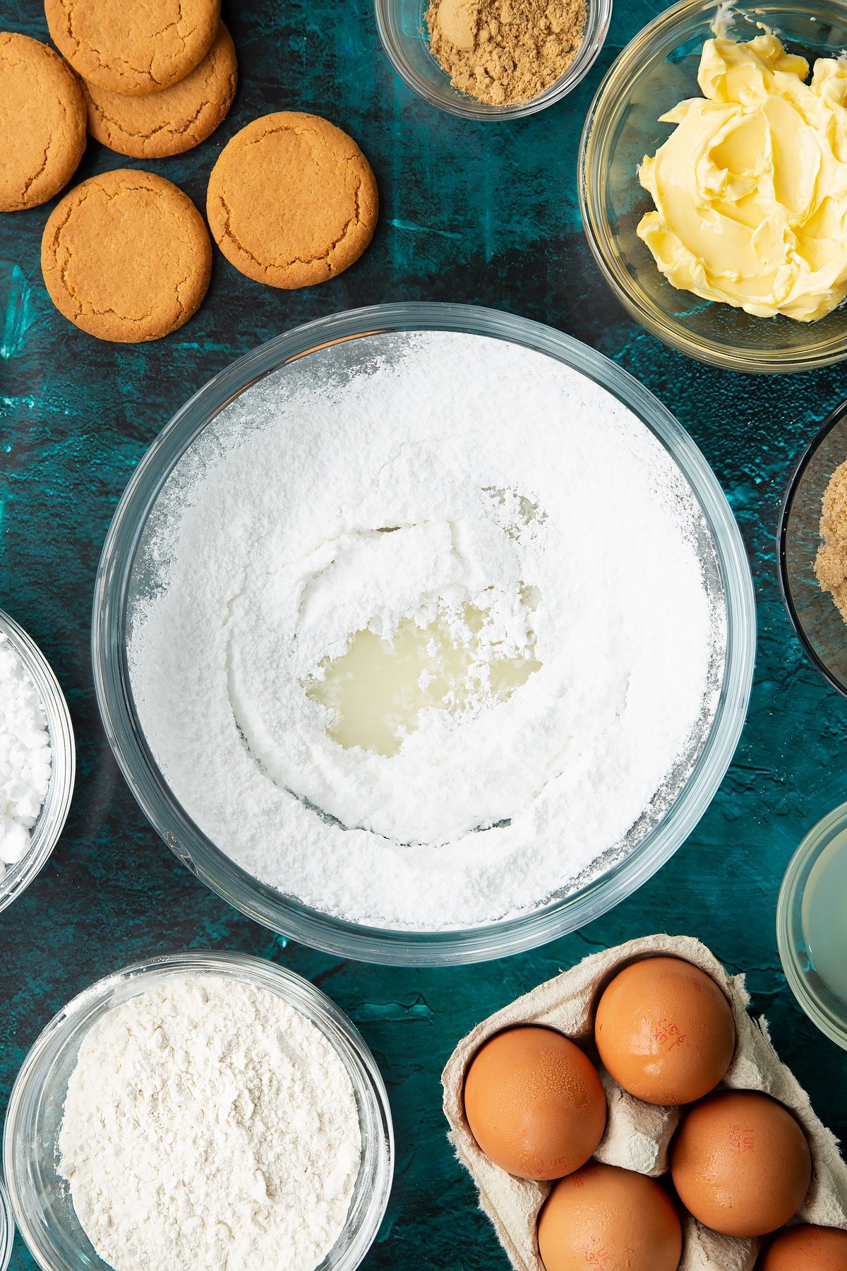 Icing sugar and lemon juice in a glass mixing bowl. Ingredients to make gingerbread donuts surround the bowl.