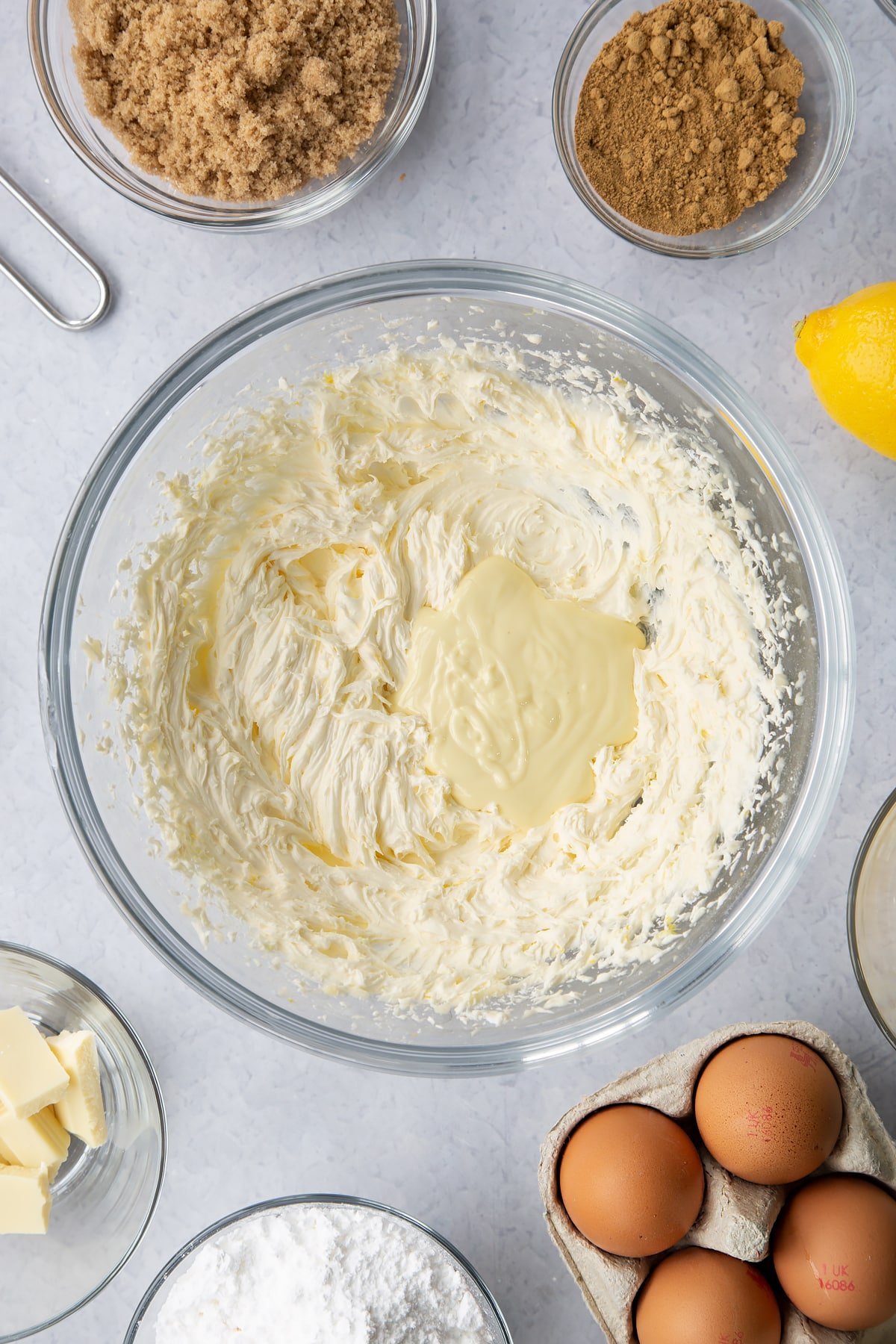 Lemon buttercream in a glass mixing bowl with white chocolate on top. Ingredients to make Gingerbread Swiss roll surround the bowl.