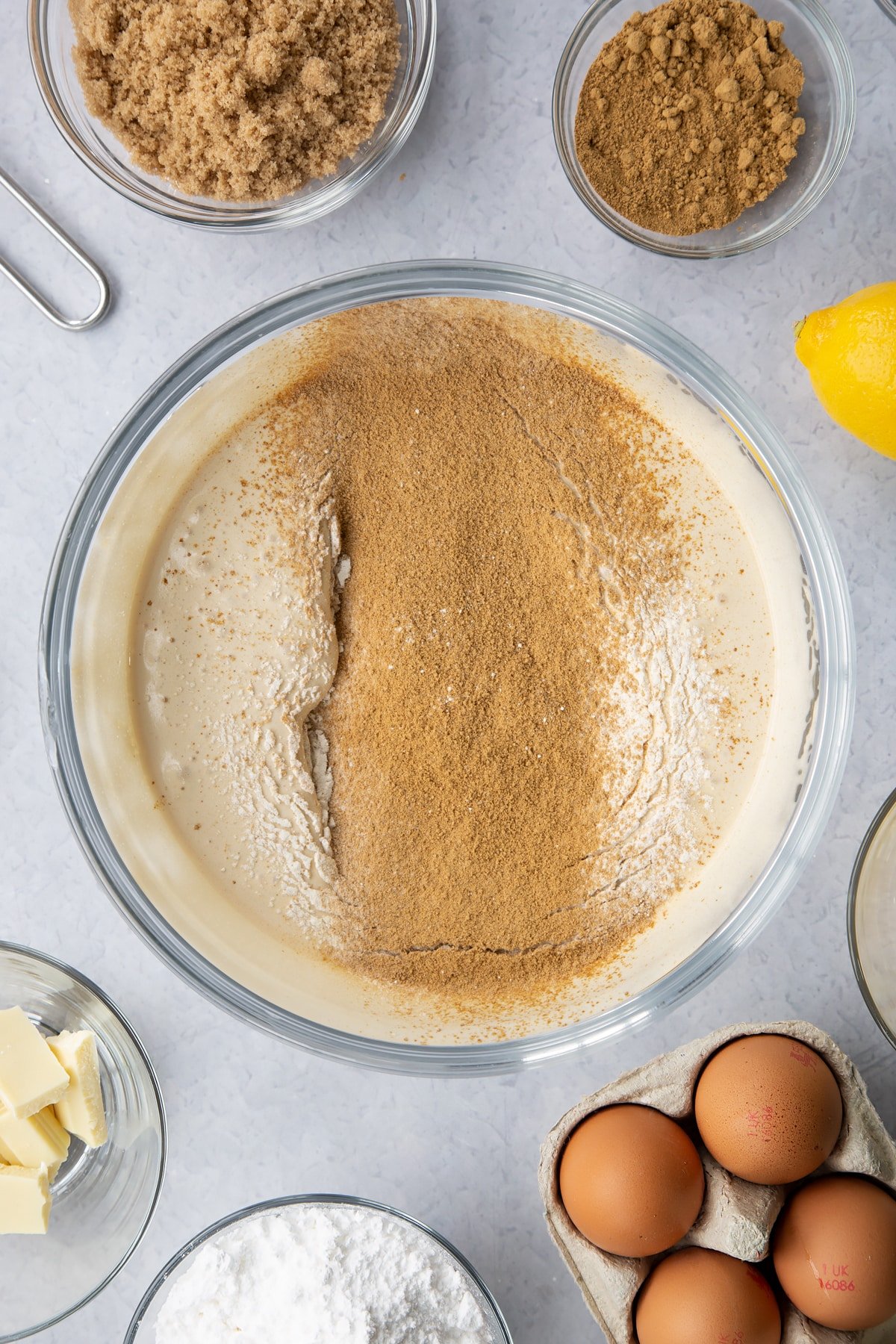 Eggs and light soft brown sugar whisked to a pale, thick mixture in a glass mixing bowl with flour and ginger on top. Ingredients to make Gingerbread Swiss roll surround the bowl.