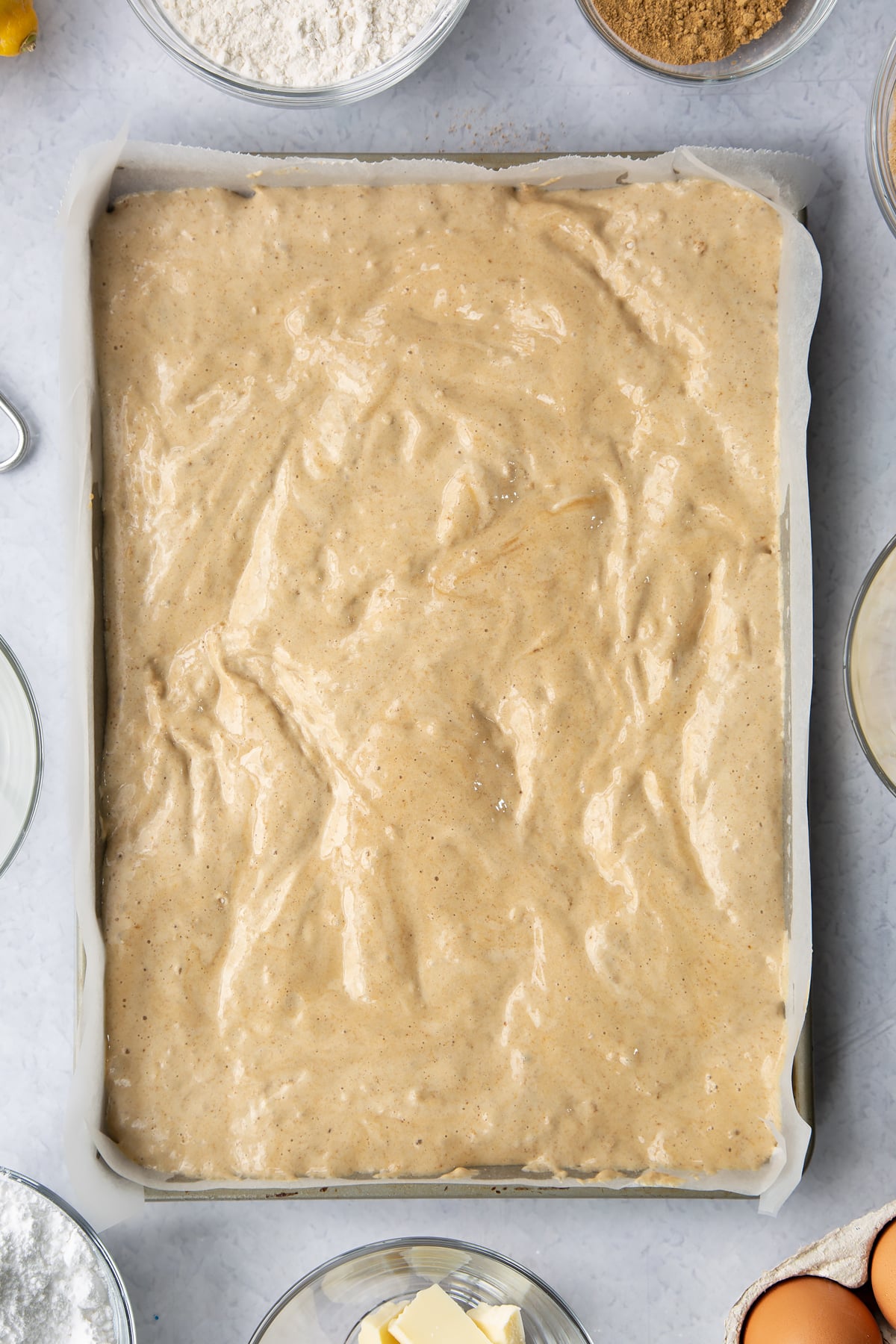 Gingerbread Swiss roll sponge batter in a lined Swiss roll tray. Ingredients to make Gingerbread Swiss roll surround the tray.