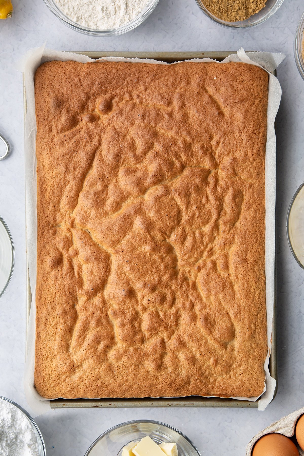 Baked gingerbread Swiss roll sponge in a lined Swiss roll tray. Ingredients to make Gingerbread Swiss roll surround the tray.