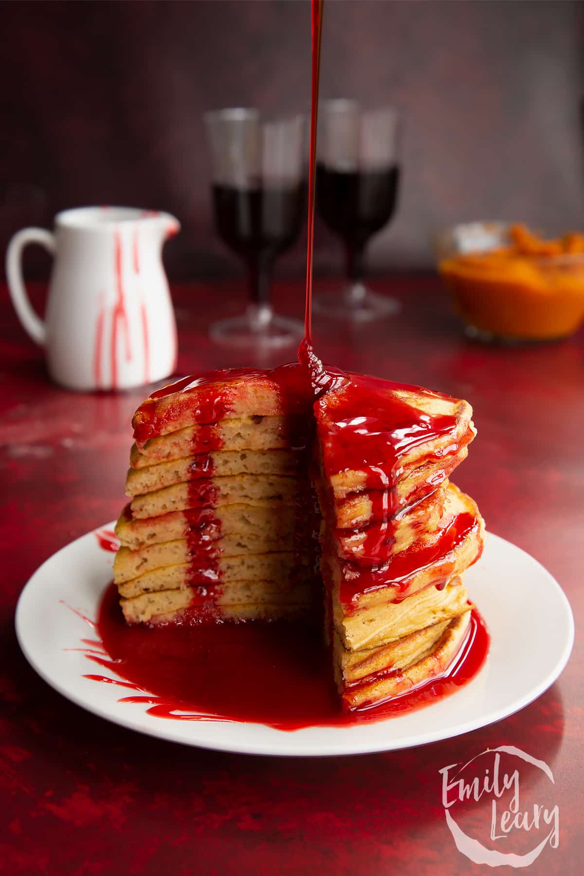 Halloween pancakes stacked on a white plate. A section has been cut away. It's being drizzled from a height with red syrup.