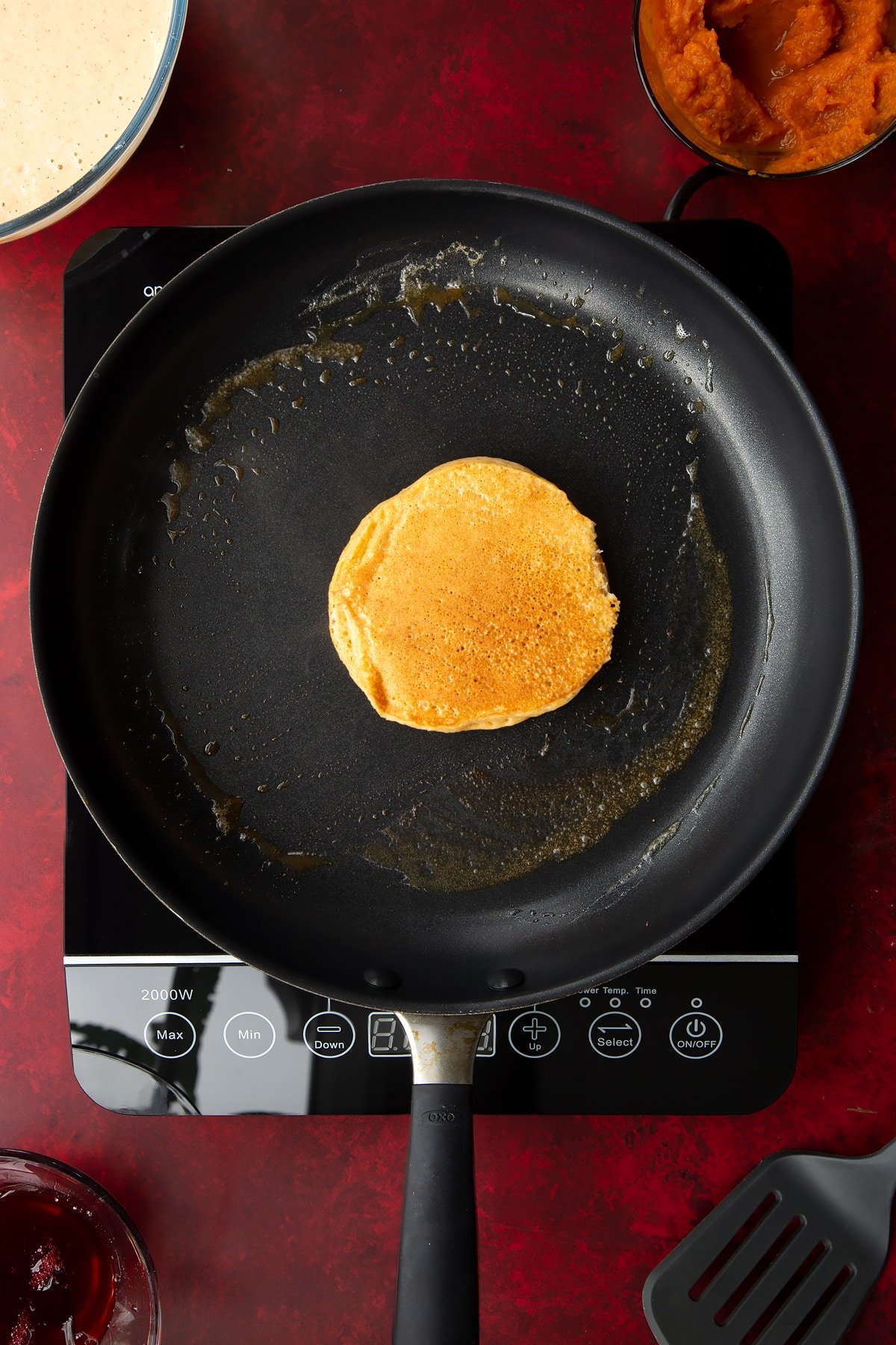 A cooked pumpkin pancake in a greased frying pan. Ingredients to make Halloween pancakes surround the pan.