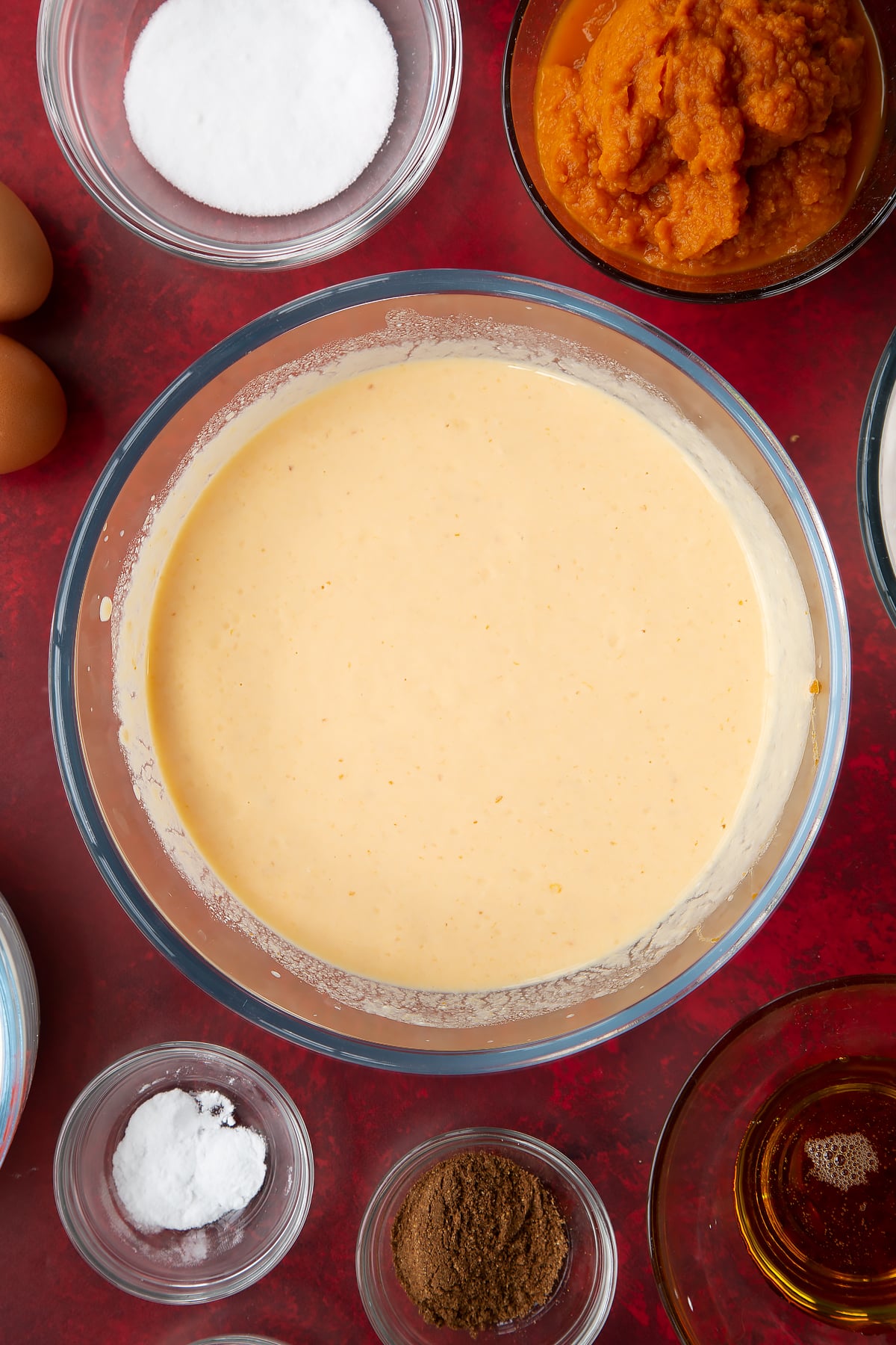 Yogurt, egg, milk and pumpkin puree, whisked together in a glass mixing bowl. Ingredients to make Halloween pancakes surround the bowl.