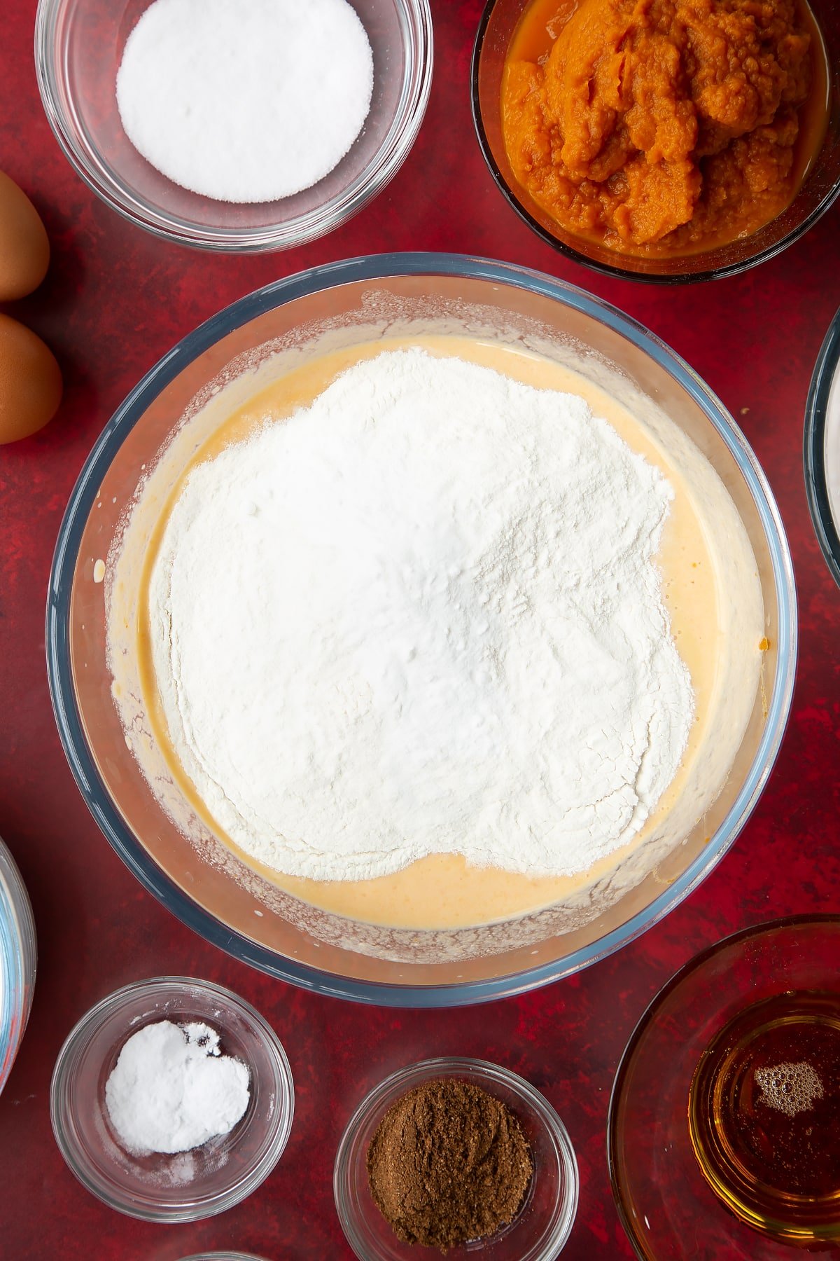 Whisked yogurt, egg, milk and pumpkin puree with flour, sugar, baking powder, bicarbonate of soda and mixed spice in a glass mixing bowl. Ingredients to make Halloween pancakes surround the bowl.