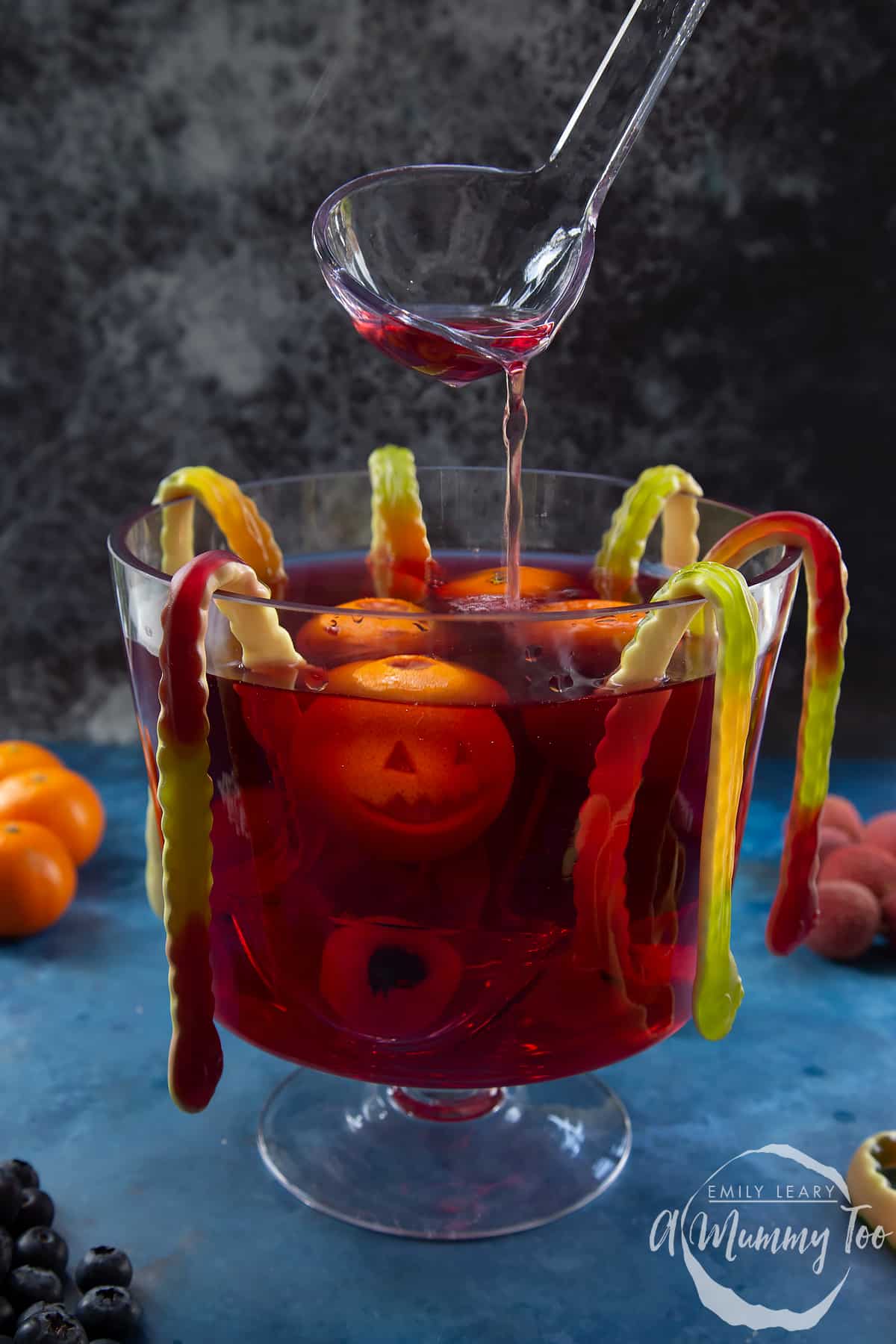 Halloween punch in a glass punch bowl. It is a red liquid, decorated with gummy snakes, jack'o'lantern clementines and lychee and blueberry eyeballs. A ladle drizzles punch into the bowl.