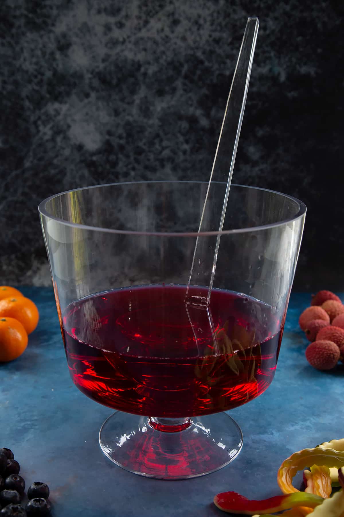 A glass punch bowl with ladle. It's part filled with cranberry juice. Ingredients to make Halloween punch surround the bowl.