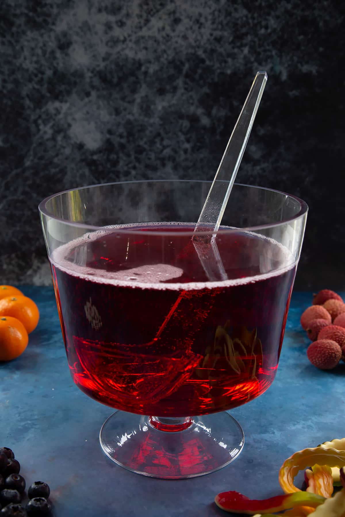 A glass punch bowl with ladle. It's filled with cranberry juice and lemonade. Ingredients to make Halloween punch surround the bowl.