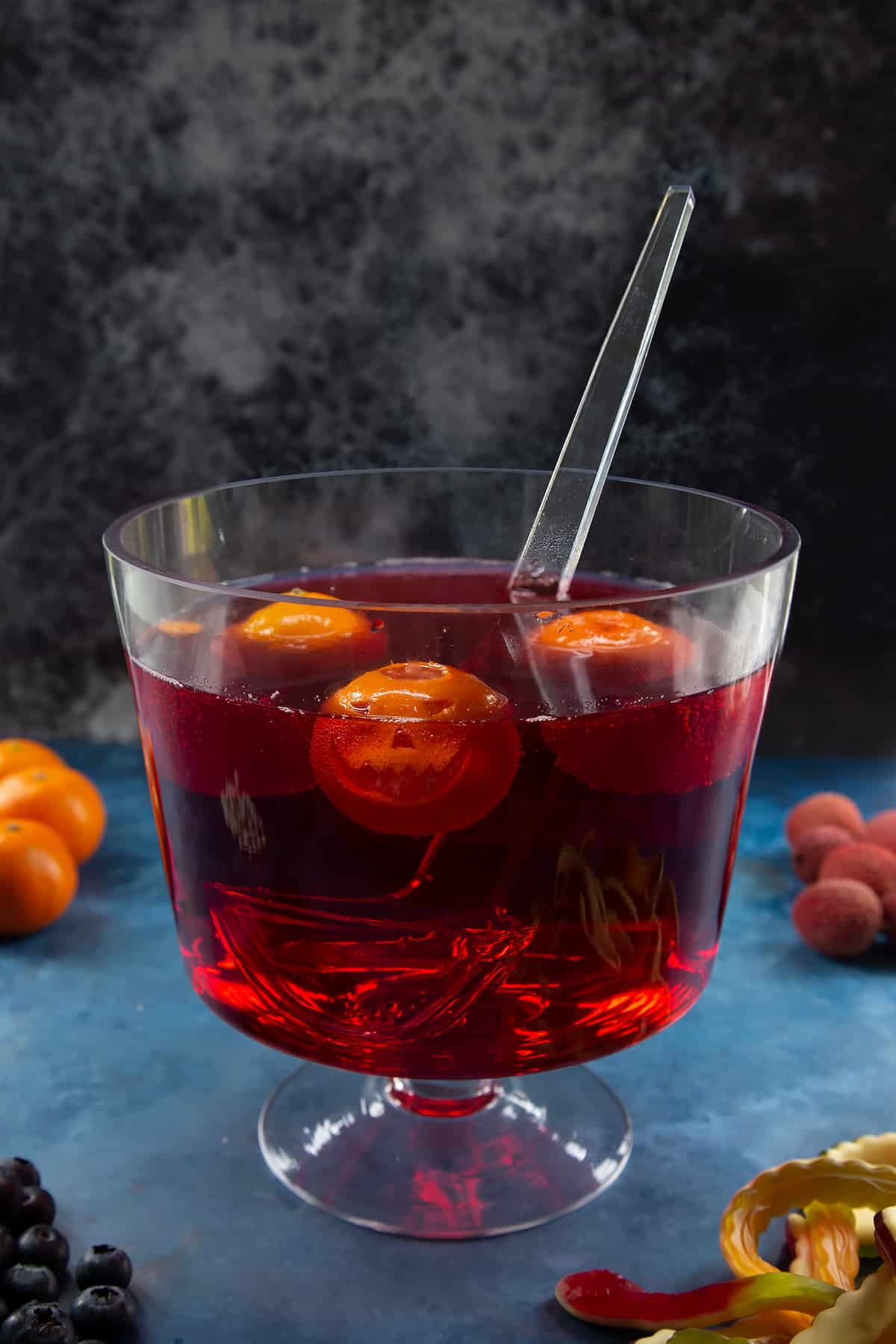 A glass punch bowl with ladle. It's filled with cranberry juice and lemonade. Jack'o'lantern clementines float in the liquid. Ingredients to make Halloween punch surround the bowl. 