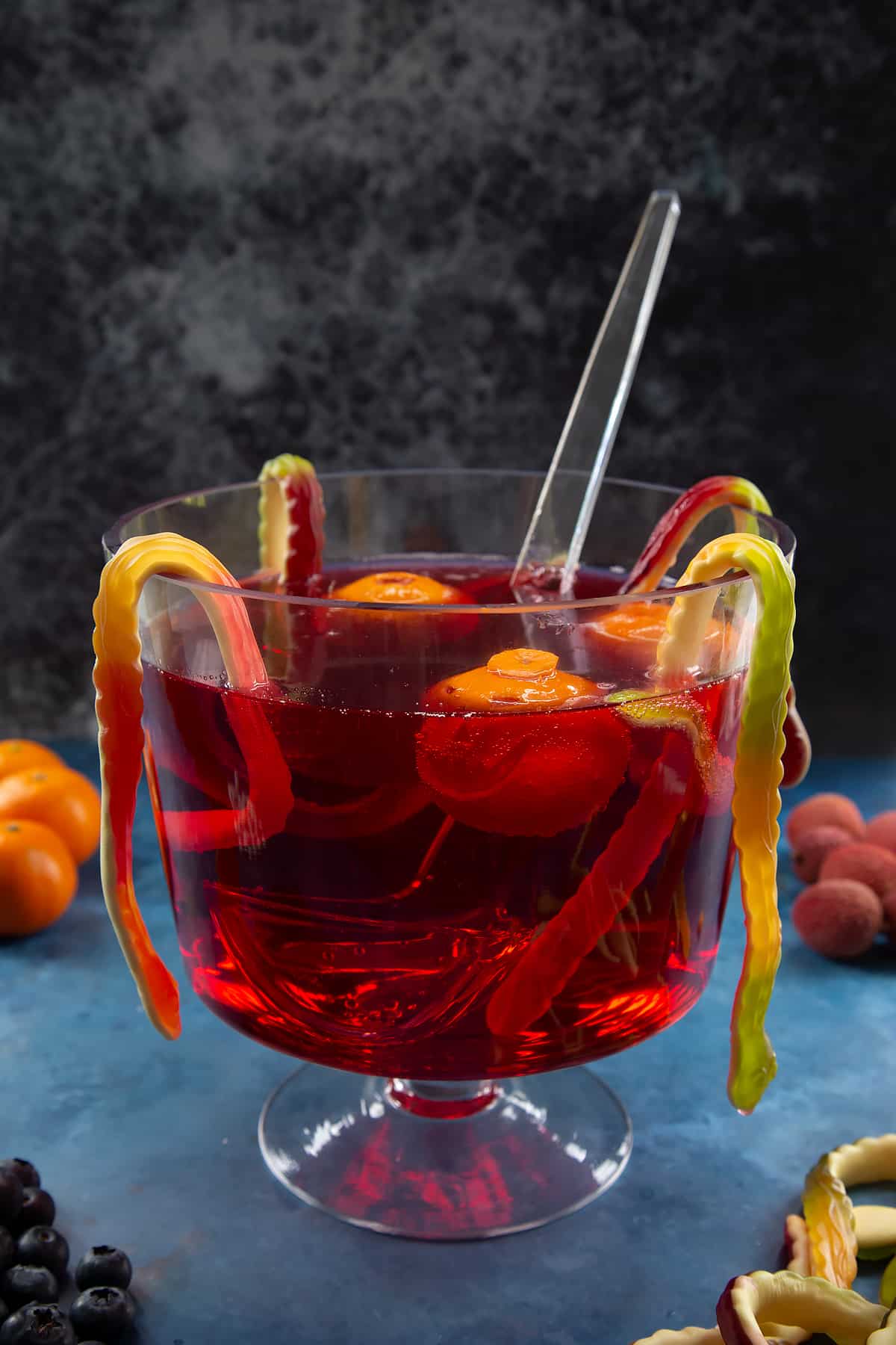 A glass punch bowl with ladle. It's filled with cranberry juice and lemonade. Jack'o'lantern clementines float in the liquid and gummy snakes hang around the bowl. Ingredients to make Halloween punch surround the bowl.