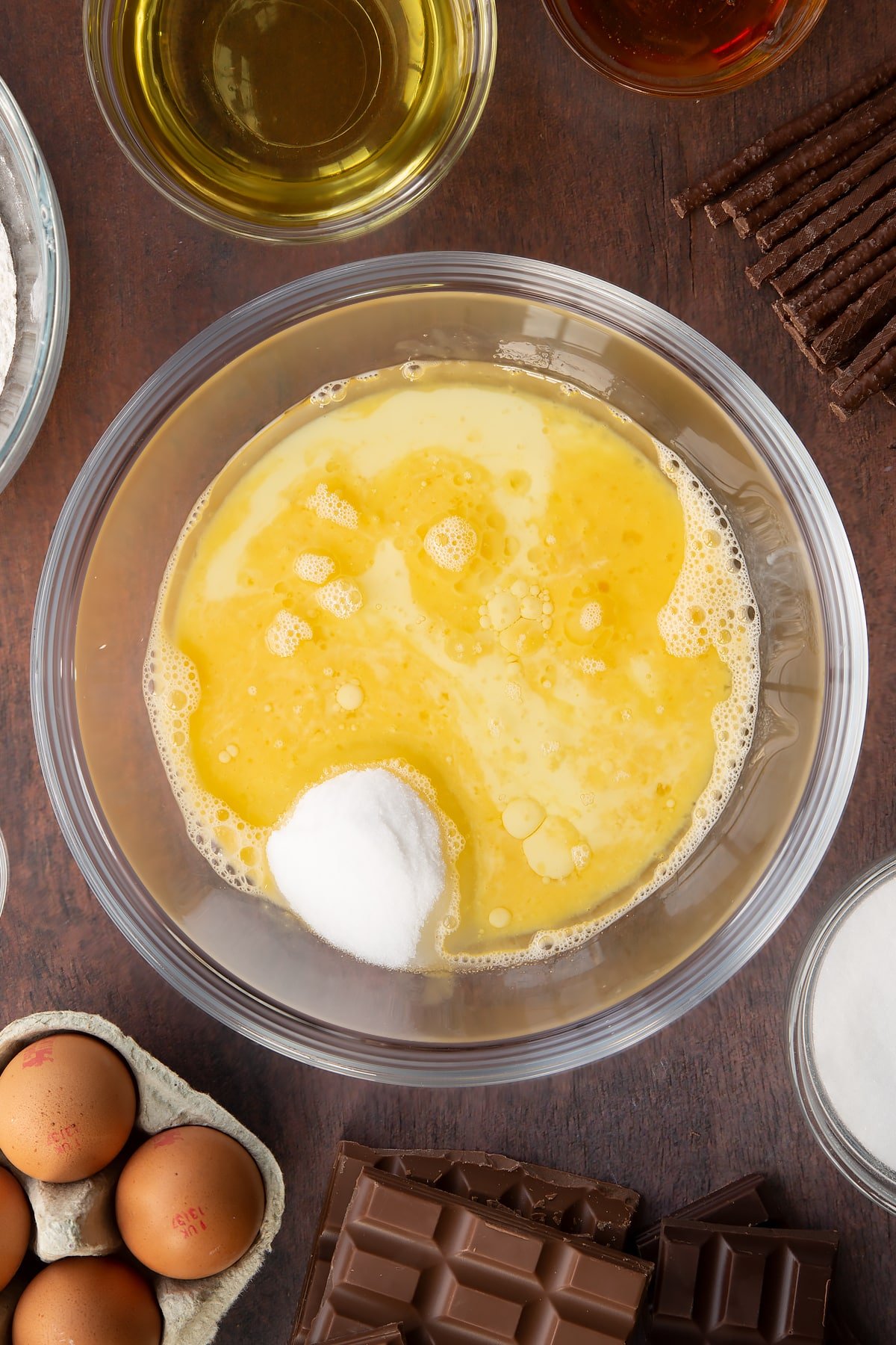 Sugar, beaten eggs, oil, milk and golden syrup in a glass mixing bowl. Ingredients to make Matchmaker cake surround the bowl. 