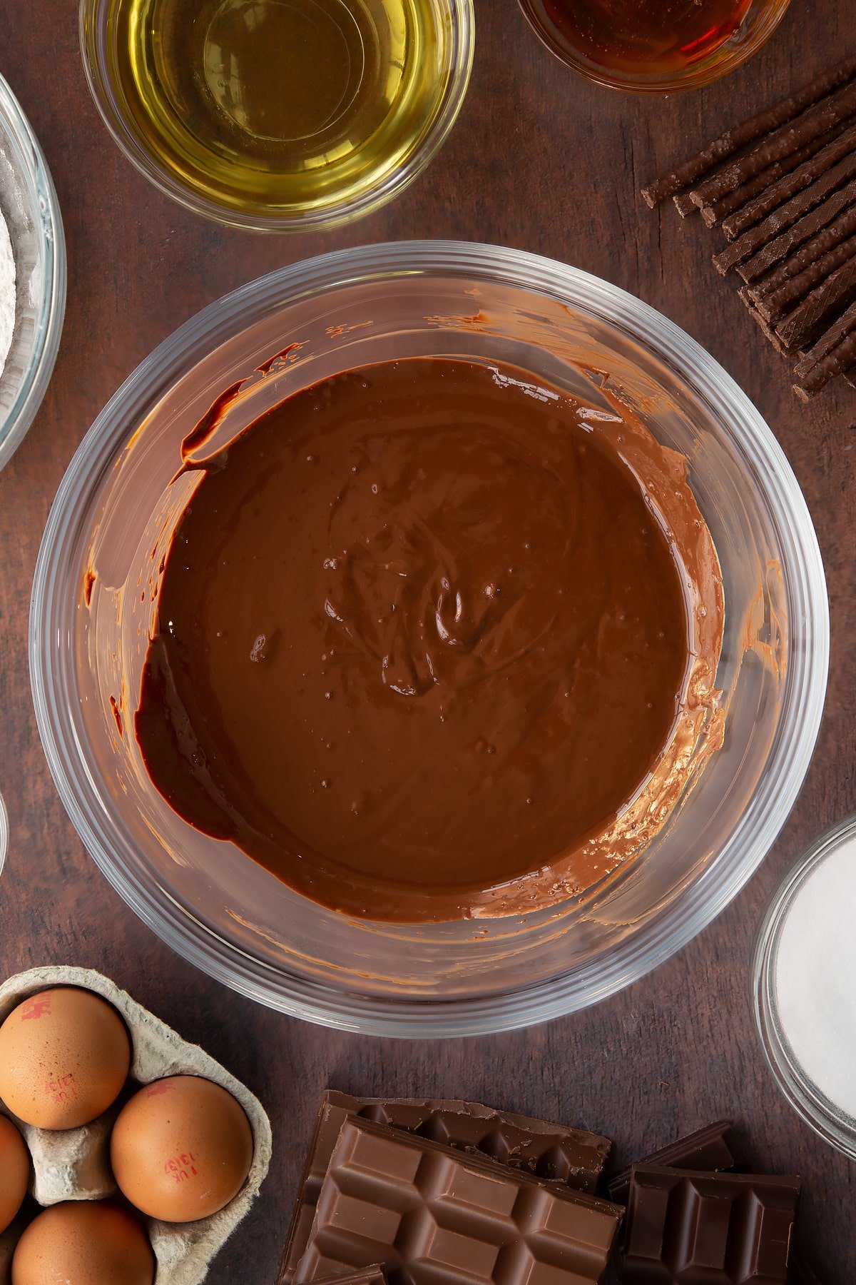 Melted dark chocolate, milk chocolate and Matchmakers in a glass mixing bowl. Ingredients to make Matchmaker cake surround the bowl. 