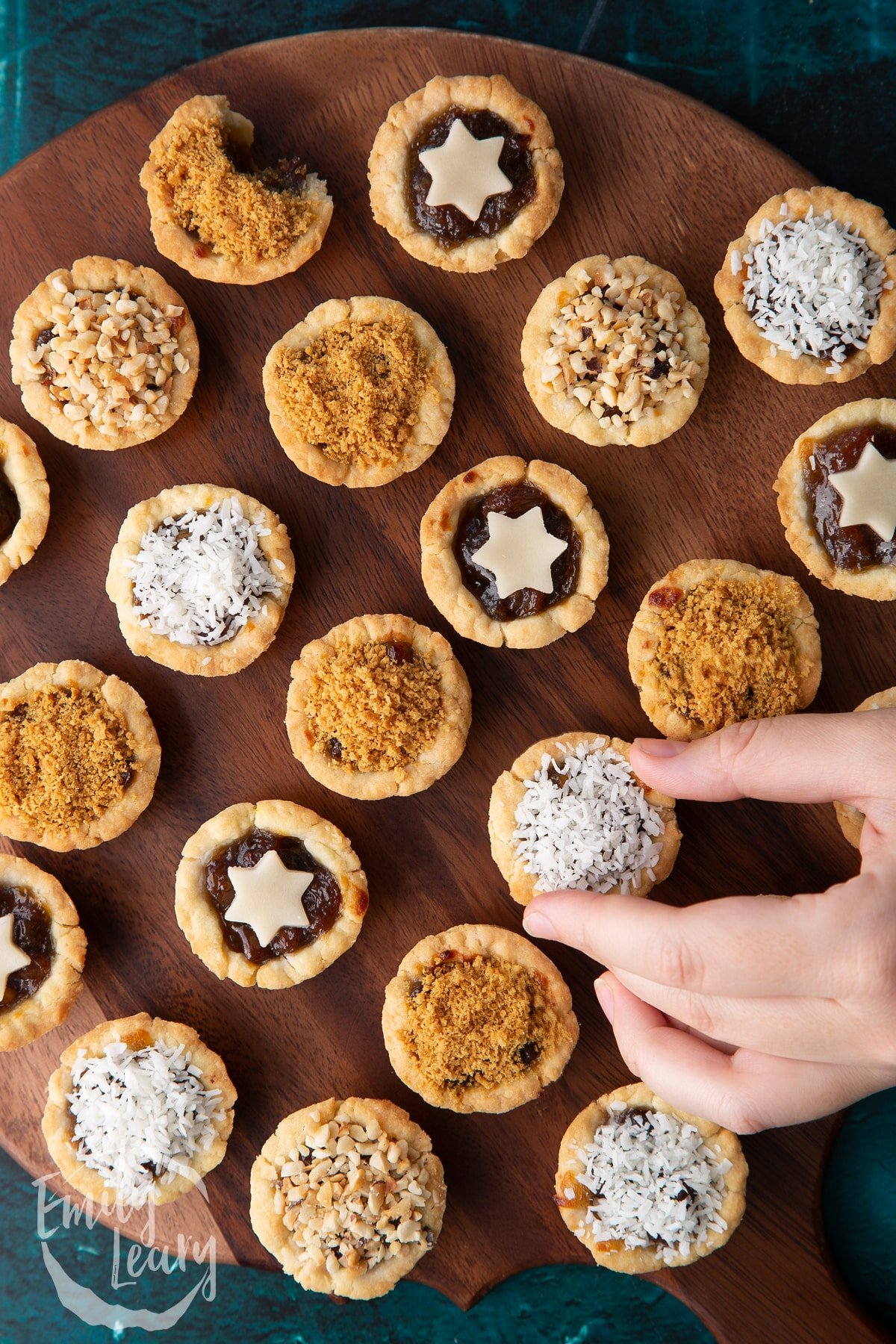 Mini mince pies, shown from above on a dark wooden board. A hand reaches for a desiccated coconut topped one.