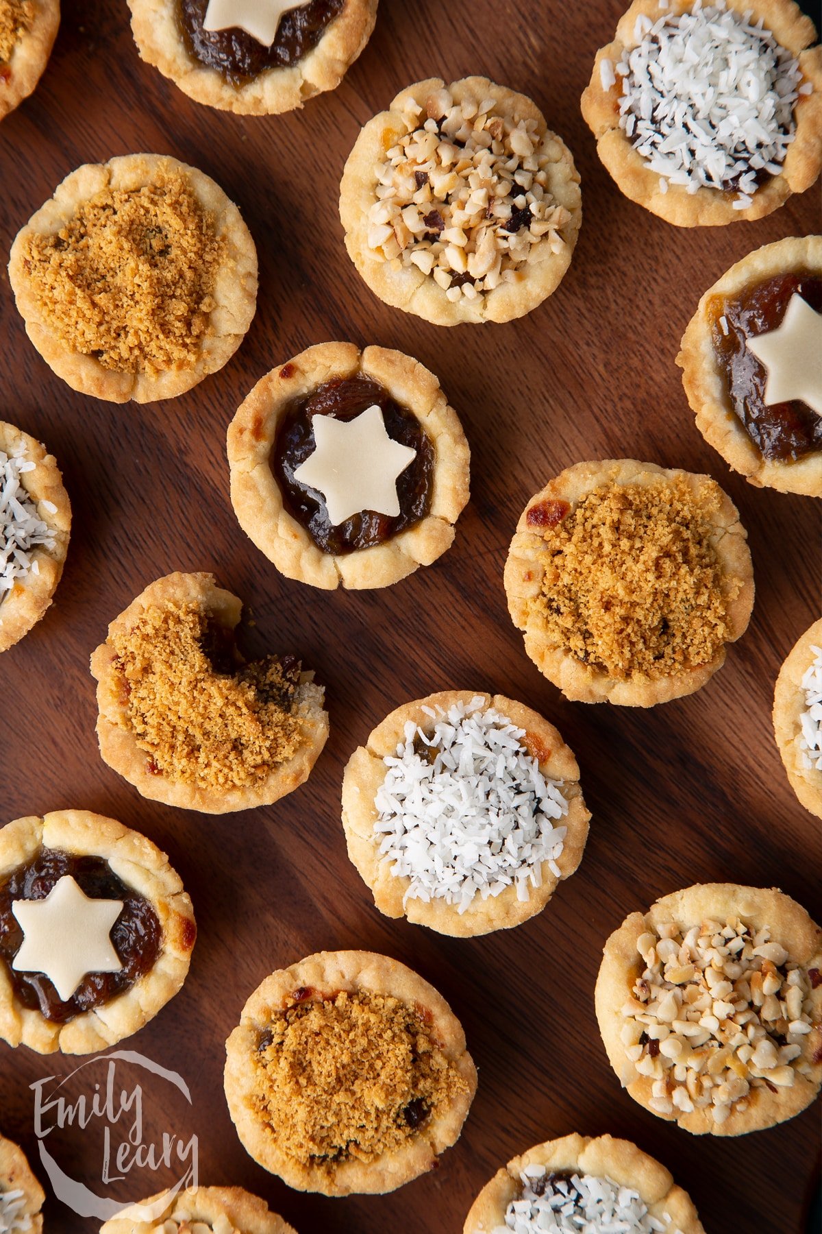 Close up on mini mince pies, shown from above on a dark wooden board. One has a bite out of it.