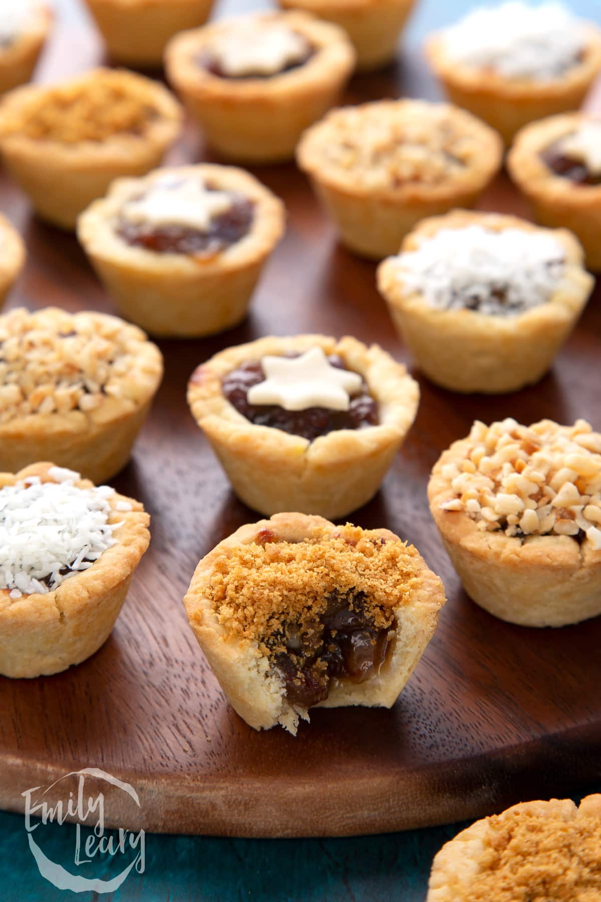 A crushed gingernut topped mini mince pie with a bite out of it. More mini mince pies are also on the dark wooden board.