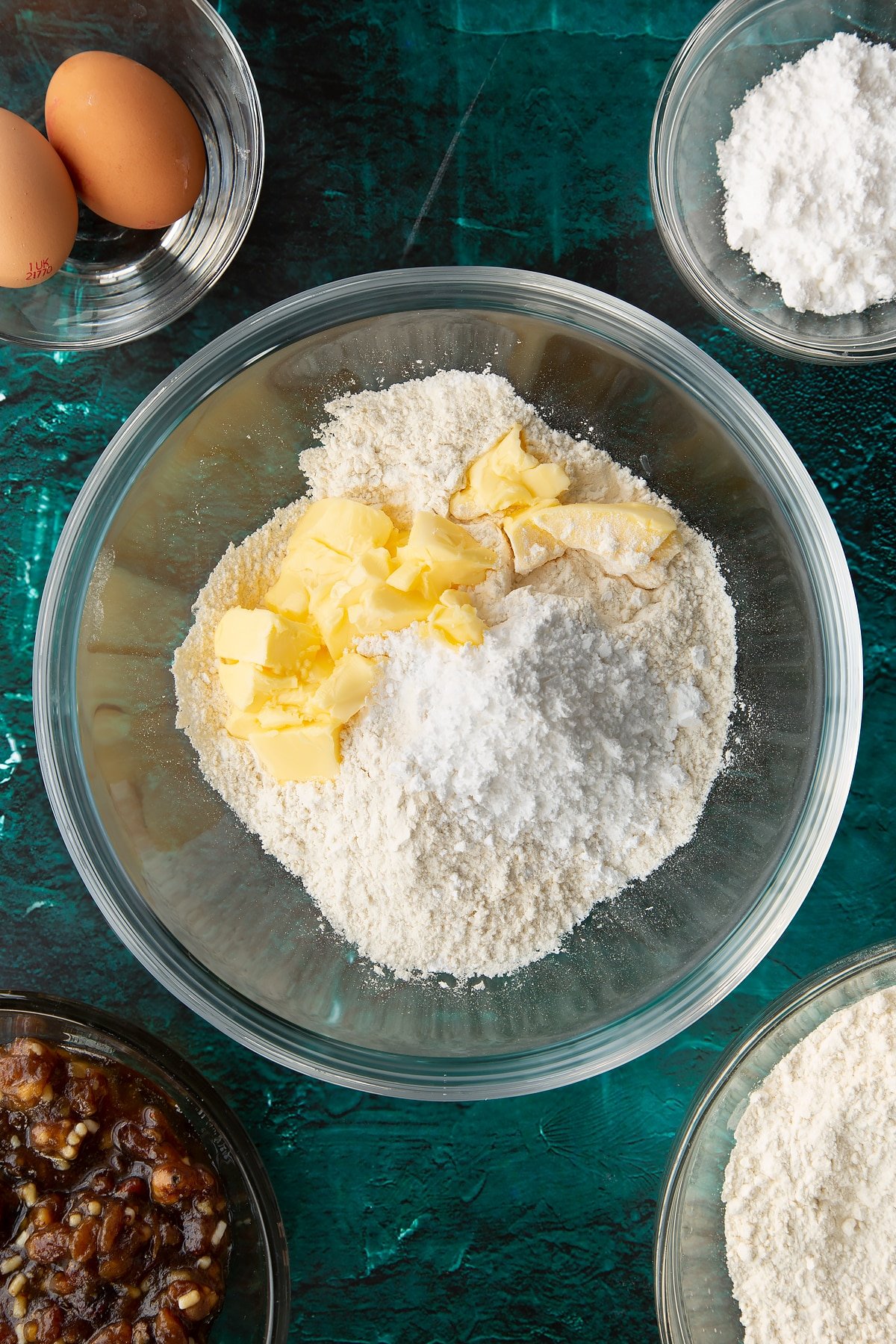 Flour, sugar and butter in a mixing bowl. Ingredients to make mini mince pies surround the bowl.