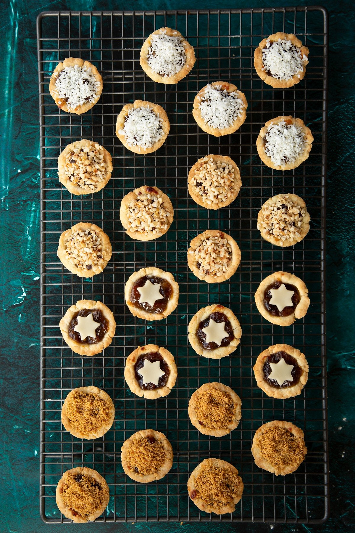 Freshly baked mini mince pies on a cooling rack. They're topped with  marzipan stars, chopped nuts, coconut or crushed gingernuts.