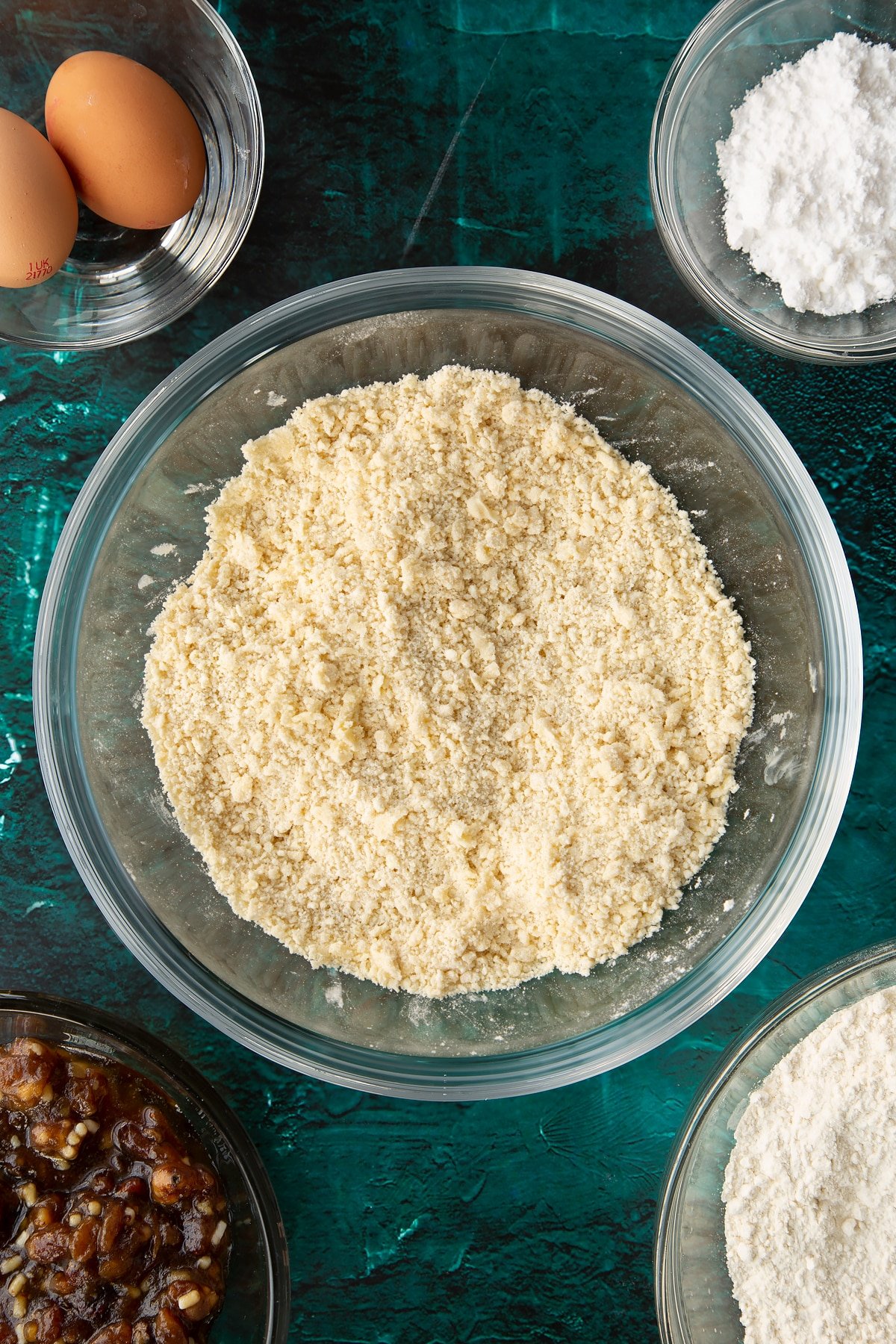 Flour, sugar and butter rubbed together in a mixing bowl. Ingredients to make mini mince pies surround the bowl.