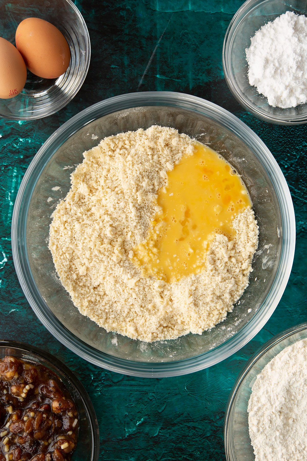 Flour, sugar and butter rubbed together in a mixing bowl with beaten egg on top. Ingredients to make mini mince pies surround the bowl.