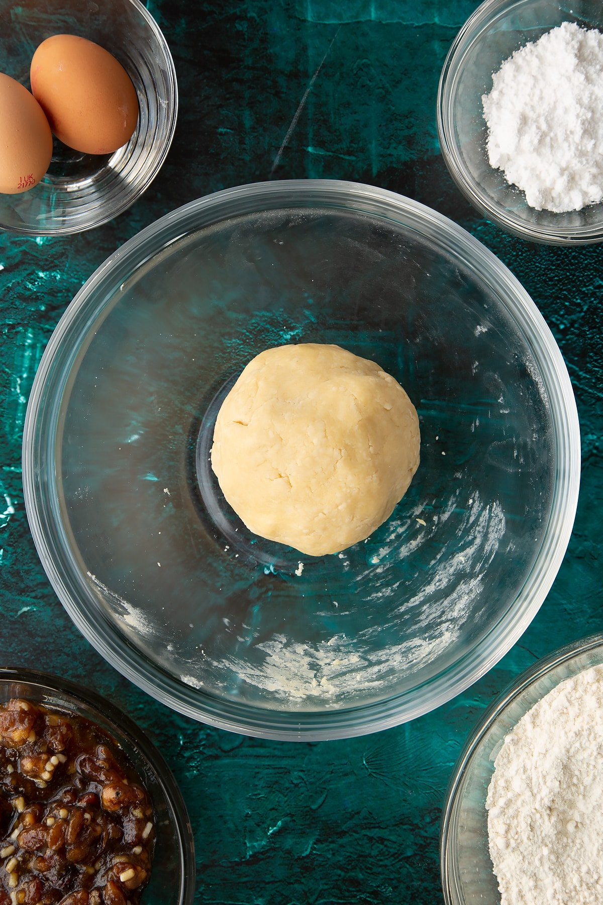 A ball of pastry in a mixing bowl. Ingredients to make mini mince pies surround the bowl.