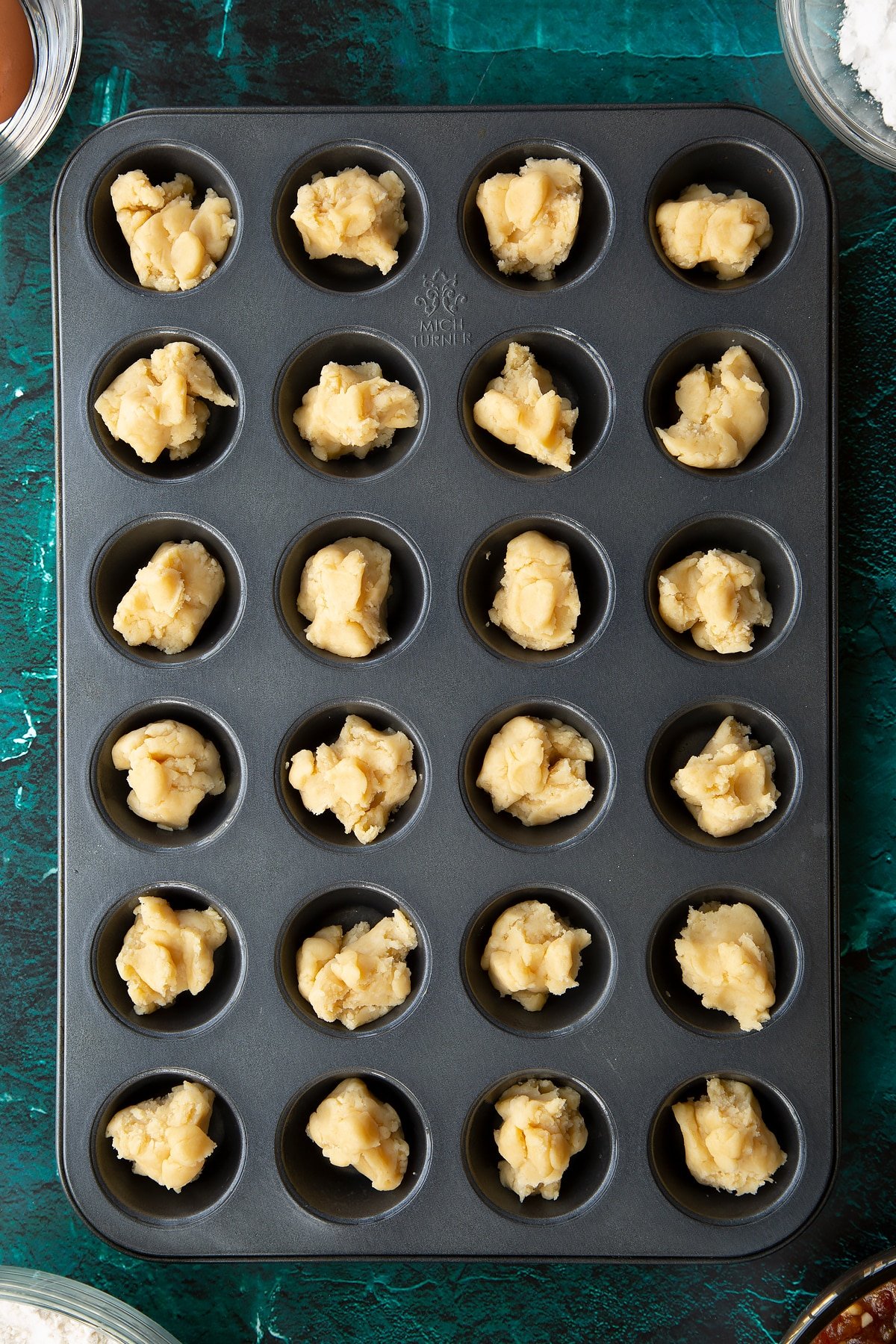 Pieces of pastry in the holes of a mini muffin tray. Ingredients to make mini mince pies surround the tray.