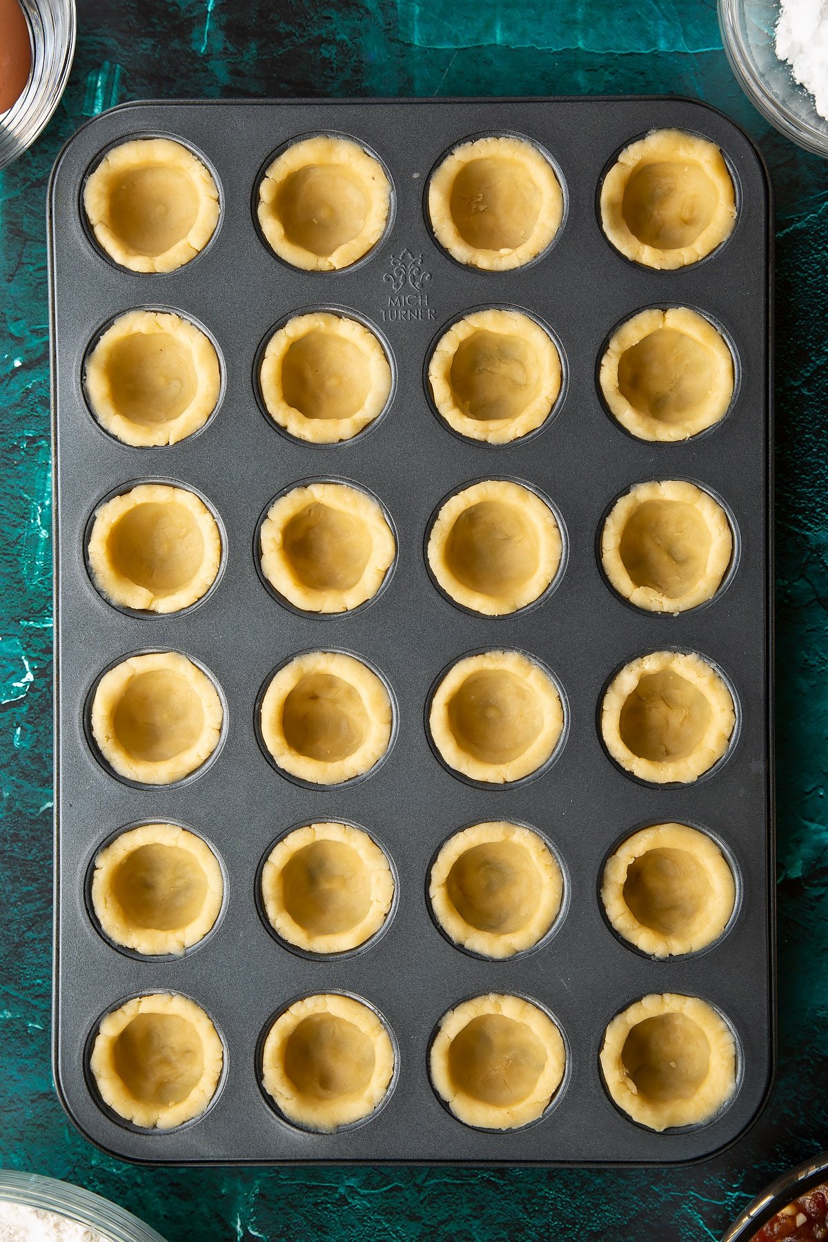 A mini muffin tray with each hole lined with pastry. Ingredients to make mini mince pies surround the tray.