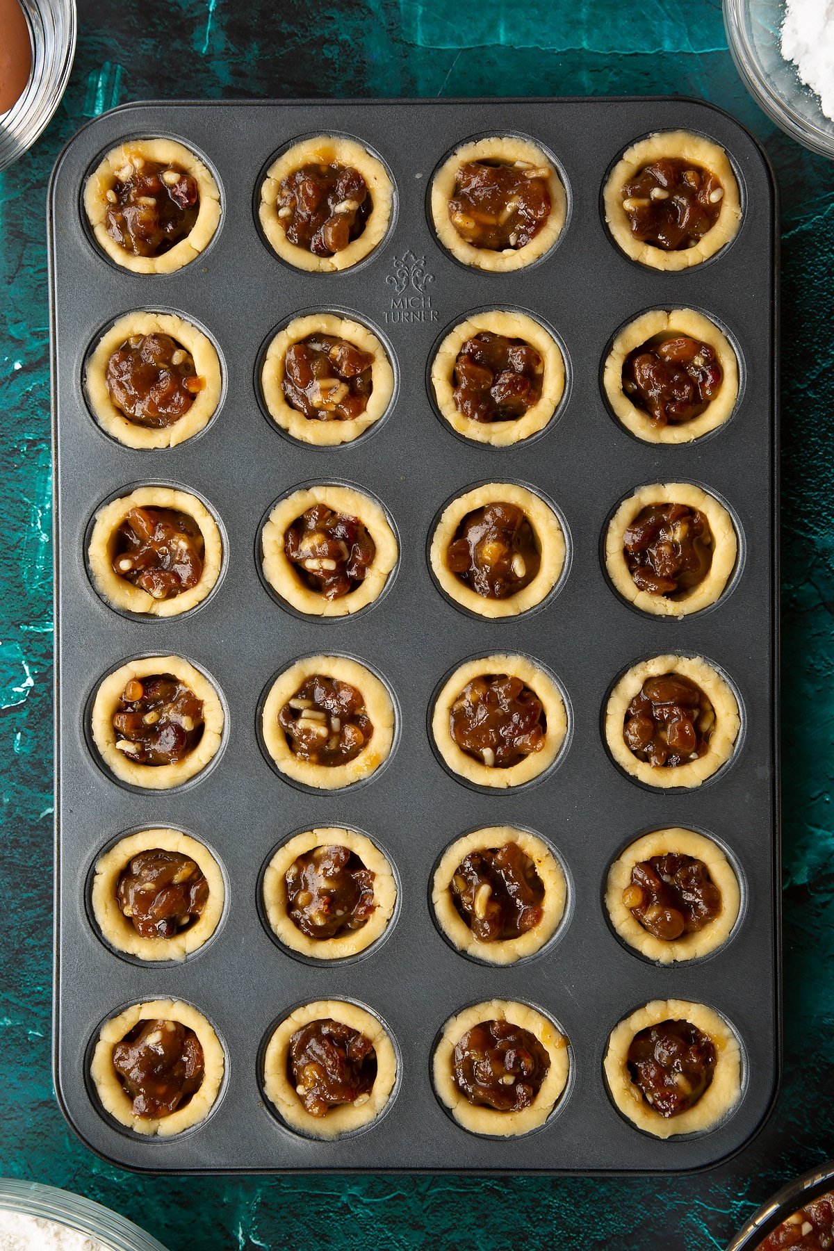 A mini muffin tray with each hole lined with pastry and filled with mincemeat. Ingredients to make mini mince pies surround the tray.