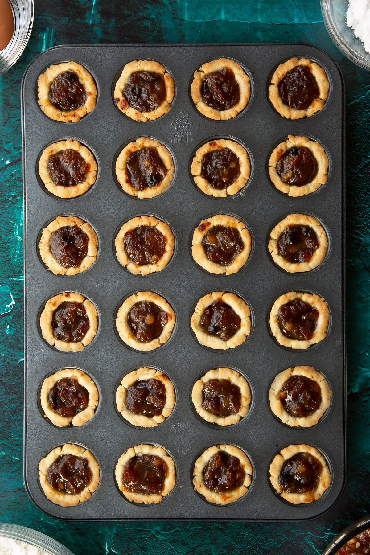 Freshly baked mini mince pies in a mini muffin tray. Ingredients to make mini mince pies surround the tray.