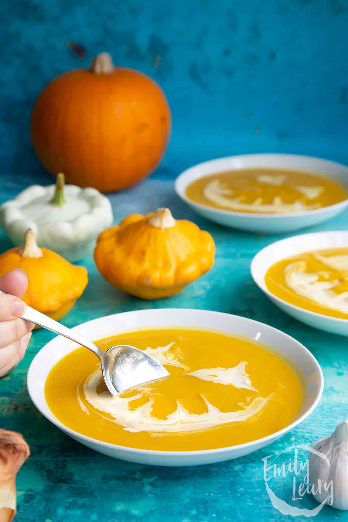 Pumpkin and sweet potato soup in a white bowl. The soup has cream swirled on top to resemble a jack'o'lantern. A hand holds a spoon, dipping into the soup.