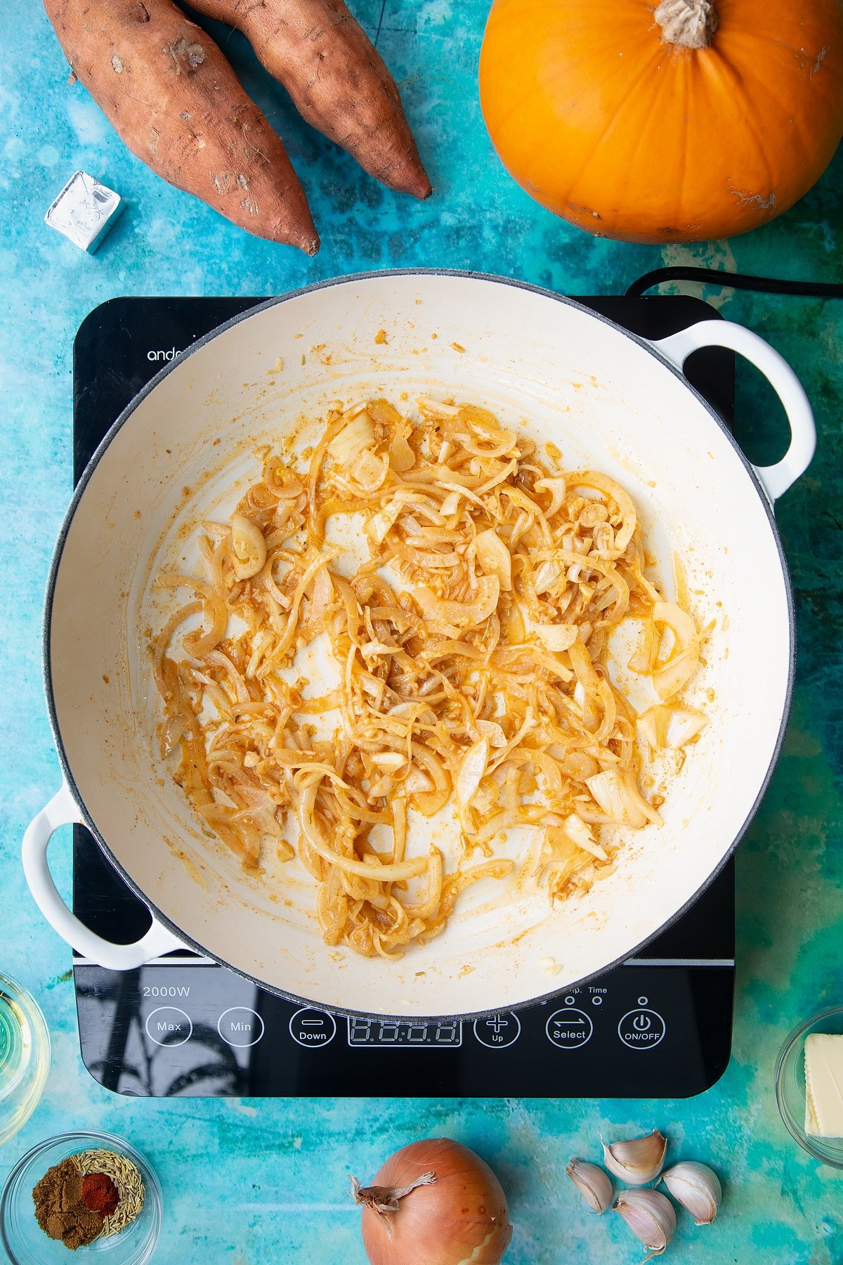 Onion, garlic, herbs and spices sweated in a large, shallow pan, from above. Ingredients to make pumpkin and sweet potato soup surround the pan.