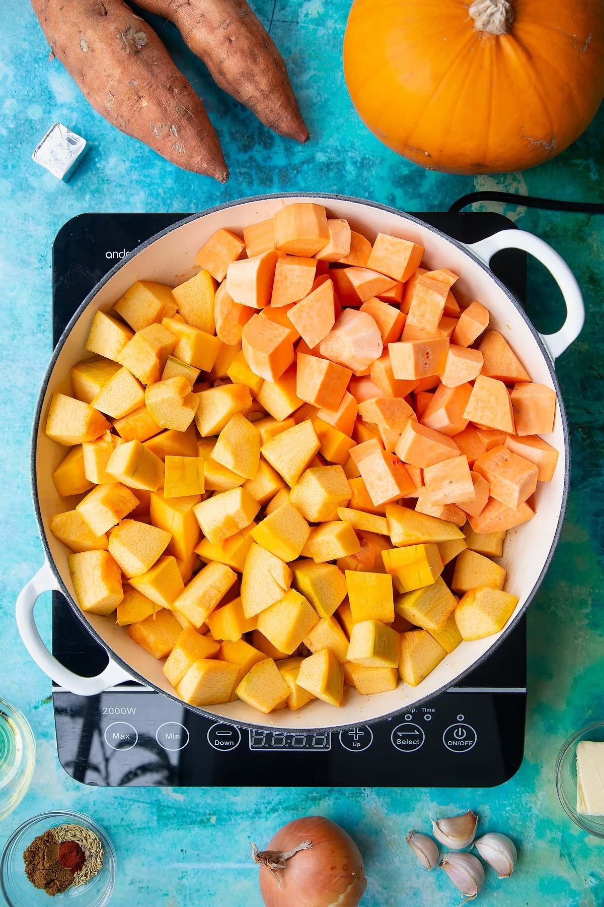 Chopped pumpkin and sweet potato on top of sweated onion and garlic in a large, shallow pan, from above. Ingredients to make pumpkin and sweet potato soup surround the pan.