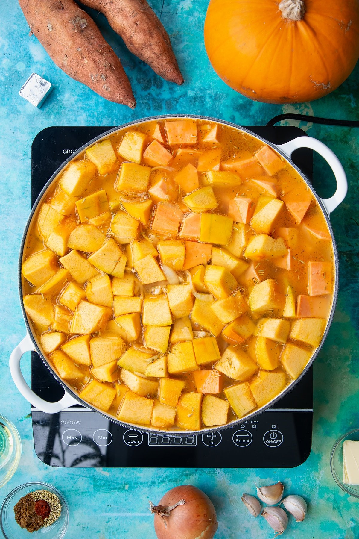 Pumpkin, sweet potato, onion, garlic and stock in a large, shallow pan, from above. Ingredients to make pumpkin and sweet potato soup surround the pan.