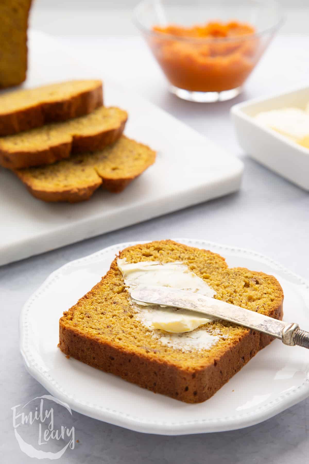 A slice of pumpkin banana bread on a small white plate. It is being buttered with a small knife.