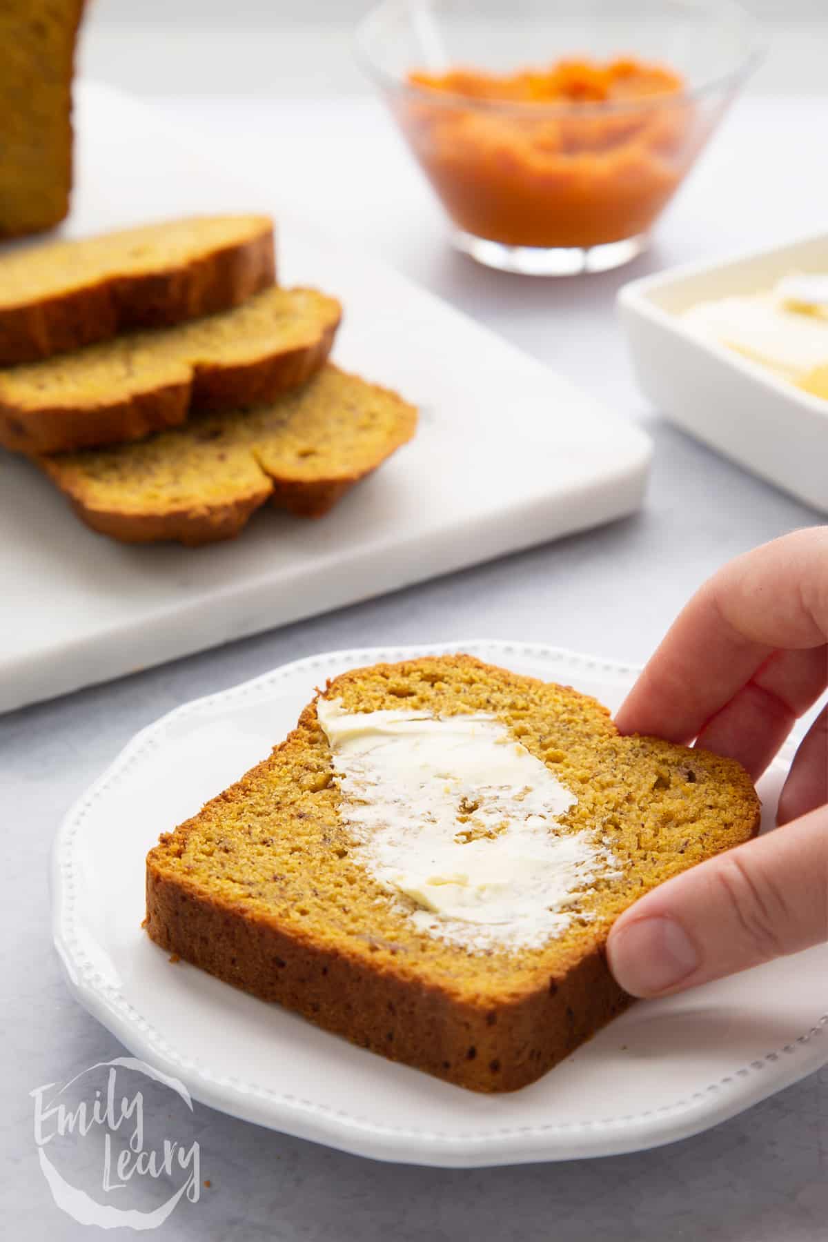 A buttered slice of pumpkin banana bread on a white plate. A hand reaches to take it.