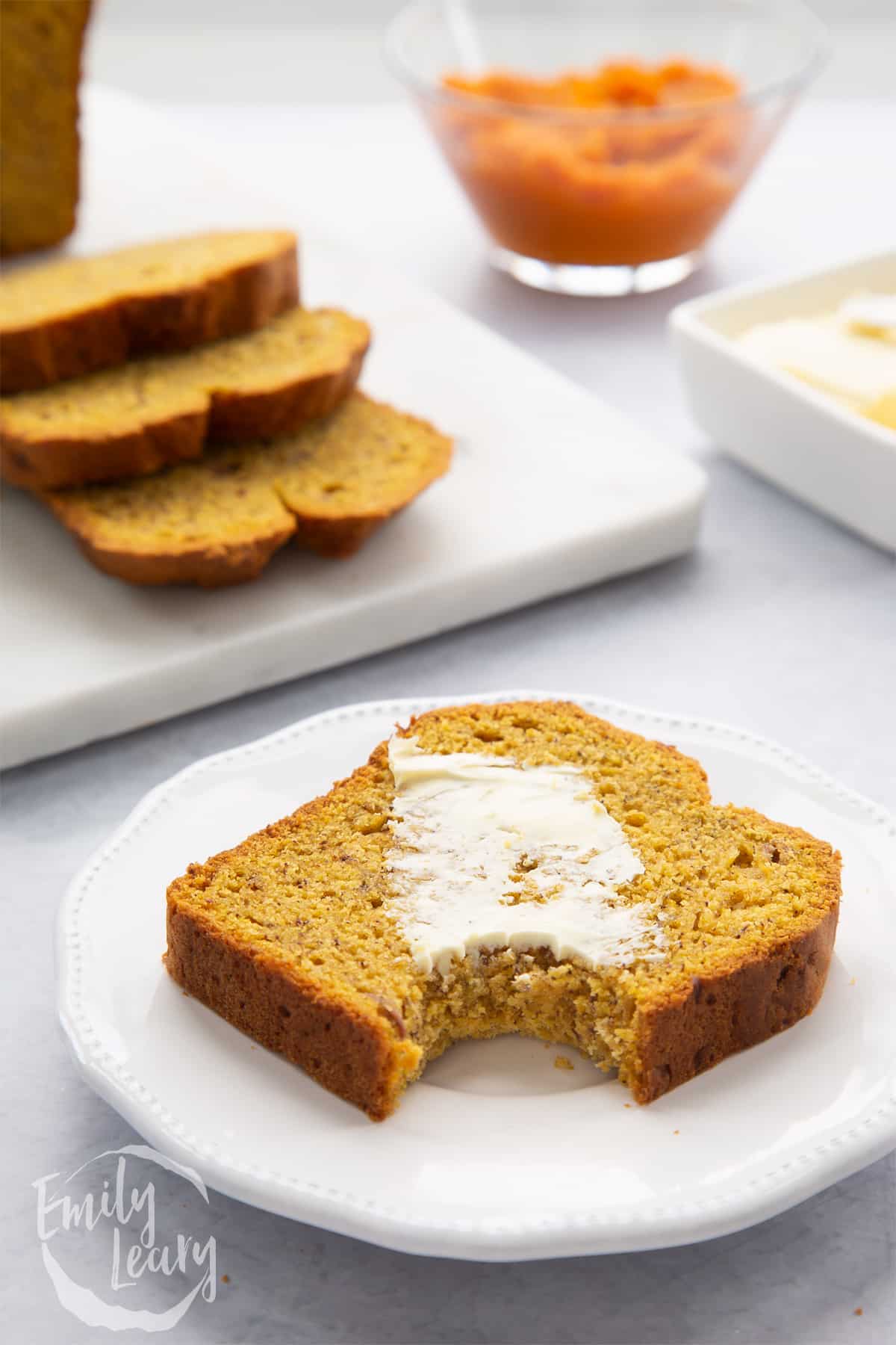 A buttered slice of pumpkin banana bread on a white plate with a bite out of it.